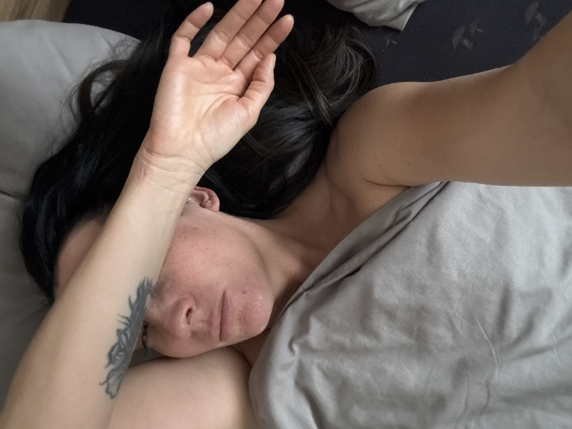 A woman is lying in bed under gray bedding. Her black hair is spread on a gray pillow. Her right arm is over her face. The lower arm has a tattoo. The woman's face is visible, including her nose, lips, and eye. The location is indiscernible from the image.