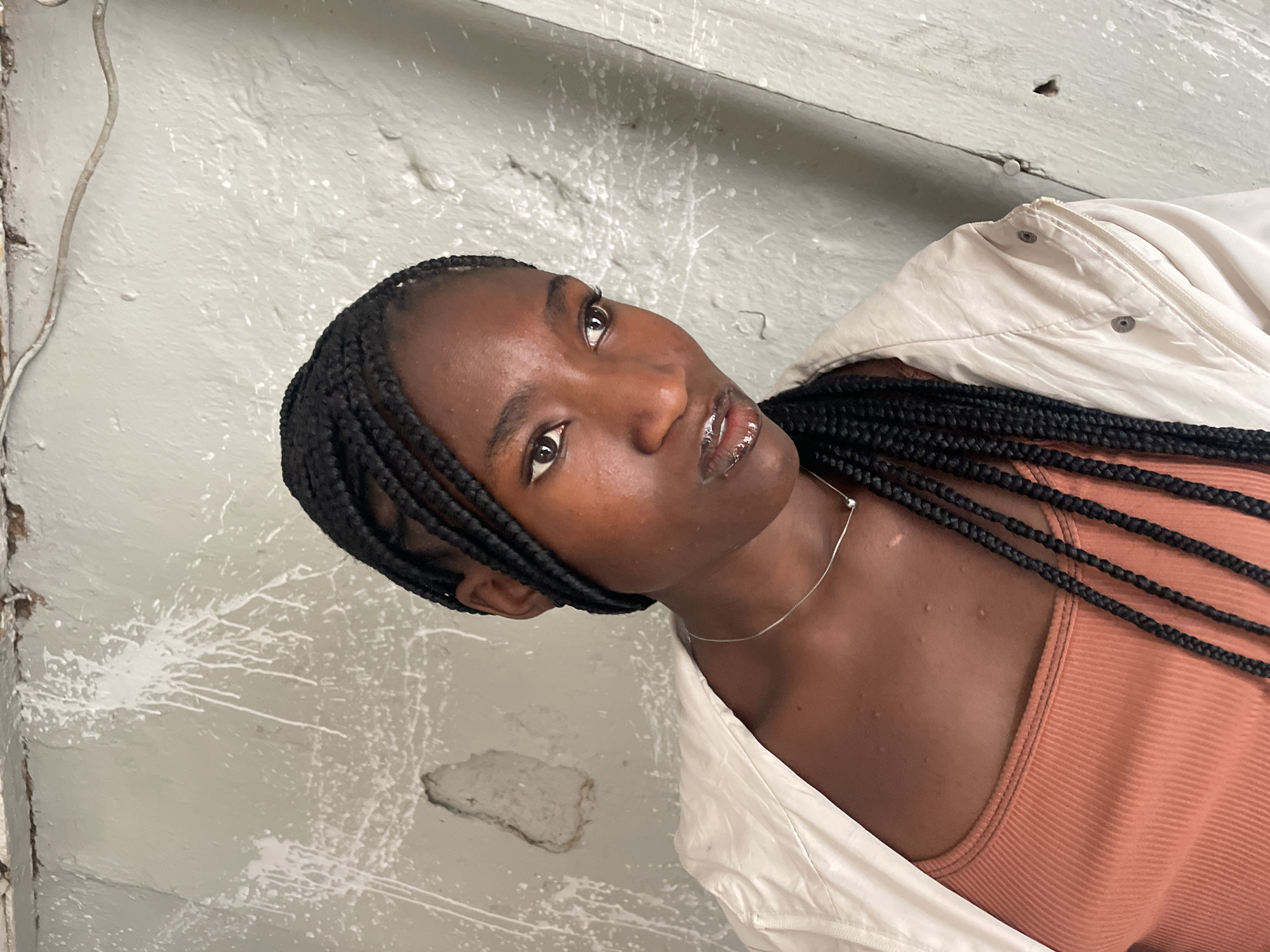 A vertical, medium shot captures a young Black woman from the chest up, set against a textured, light gray wall. The image is contextualized to be in Jos, Nigeria. She has dark skin and her hair is styled in long, black box braids, which fall over her left shoulder. She is wearing a light-colored, possibly off-white, jacket with a zipper and snap buttons, layered over an orange ribbed tank top. A delicate silver chain necklace is visible around her neck.
Her head is tilted slightly to her right, and she looks upwards and to the viewer's right with a contemplative or serene expression. Her lips are slightly parted and appear glossy, reflecting light. The background is a rough, light gray wall, likely concrete or painted plaster, adorned with numerous white paint splatters and drips. A thin, light-colored wire or cable runs vertically along the wall on the left side of the frame. There are also visible imperfections and areas of damaged plaster on the wall's surface. No visible text is present in the image.