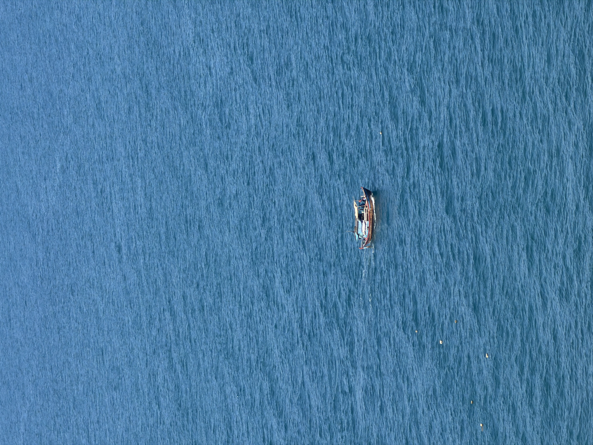 An aerial, medium-angle, full shot captures a single, small, wooden boat navigating across the vast expanse of the deep blue ocean off the coast of Nam Nha Trang Ward, Vietnam. The boat, oriented slightly to the right, has a covered cabin area and appears to have at least two individuals visible on deck. The water is choppy, with numerous small waves creating a textured surface. Several small, white objects, possibly buoys or debris, are scattered across the ocean's surface, some in the foreground and others near the boat. The image conveys a sense of isolation and the immense scale of the ocean surrounding the solitary vessel.