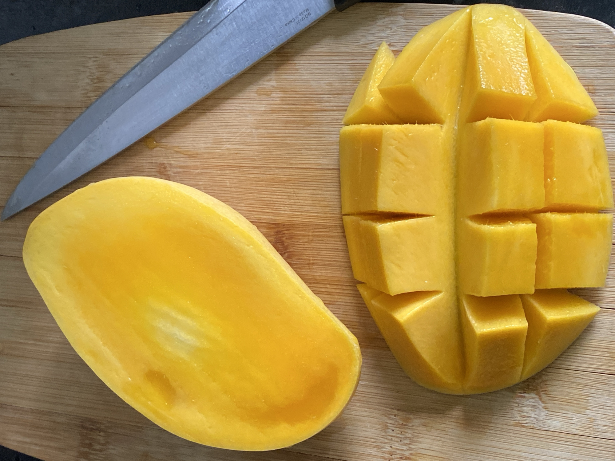 A mango cut in the hedgehog style sits next to the other half of the mango and a knife on a cutting board. The city of Chaikovsky, Russia, is not discernible from the given image.