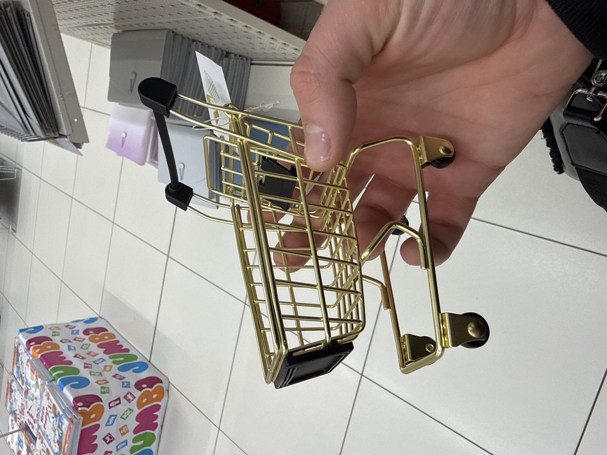 A person's hand holds a miniature, gold-colored shopping cart in an indoor setting, possibly a store. The background shows shelves with various products, including packaged items, along with a colorful box labeled "Jumbo" on the floor. The floor is covered in white square tiles with gray grout lines. A black strap is visible on the person's shoulder. The location is Stara Zagora, Bulgaria, inferred from the presence of Jumbo (a popular toy store) in the scene.