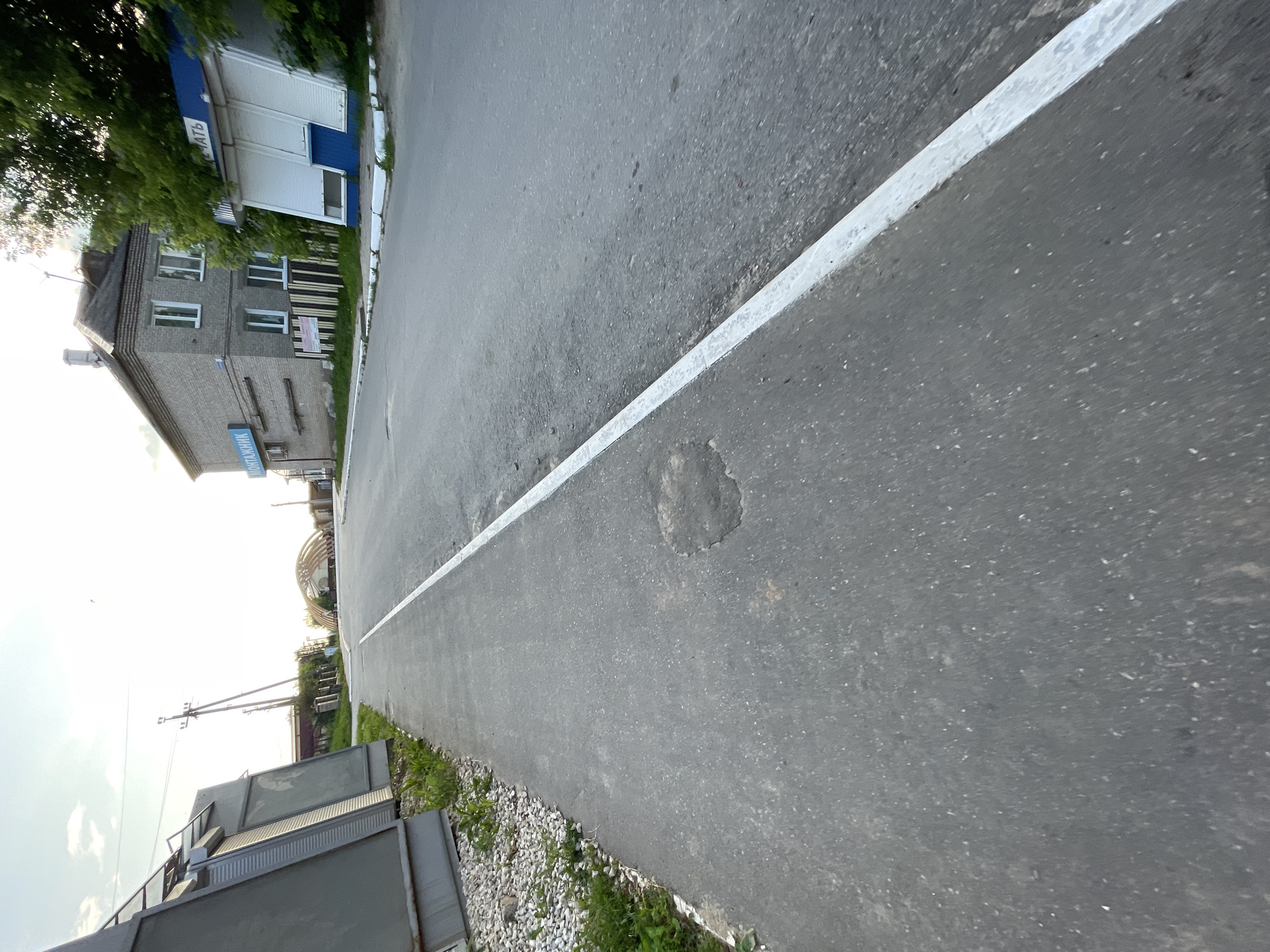 The image shows a paved road in the town of Krasnye Baki, Russia. On the left side of the road is a sidewalk, bordered by gravel and some sparse green vegetation. Further left, a tall, gray, angular structure is visible. Across the road, there is a brick building with a dark roof and multiple windows. To the right of this building is a smaller structure with a blue and white facade, featuring white rolling shutters. Above the road, power lines stretch across the scene, supported by utility poles. In the distance, an arched structure with a roof is visible, suggesting a public space or building. The sky above is bright and appears to be daytime, with a few wispy clouds. The overall impression is of a quiet, possibly rural or suburban, street scene in a Russian town.