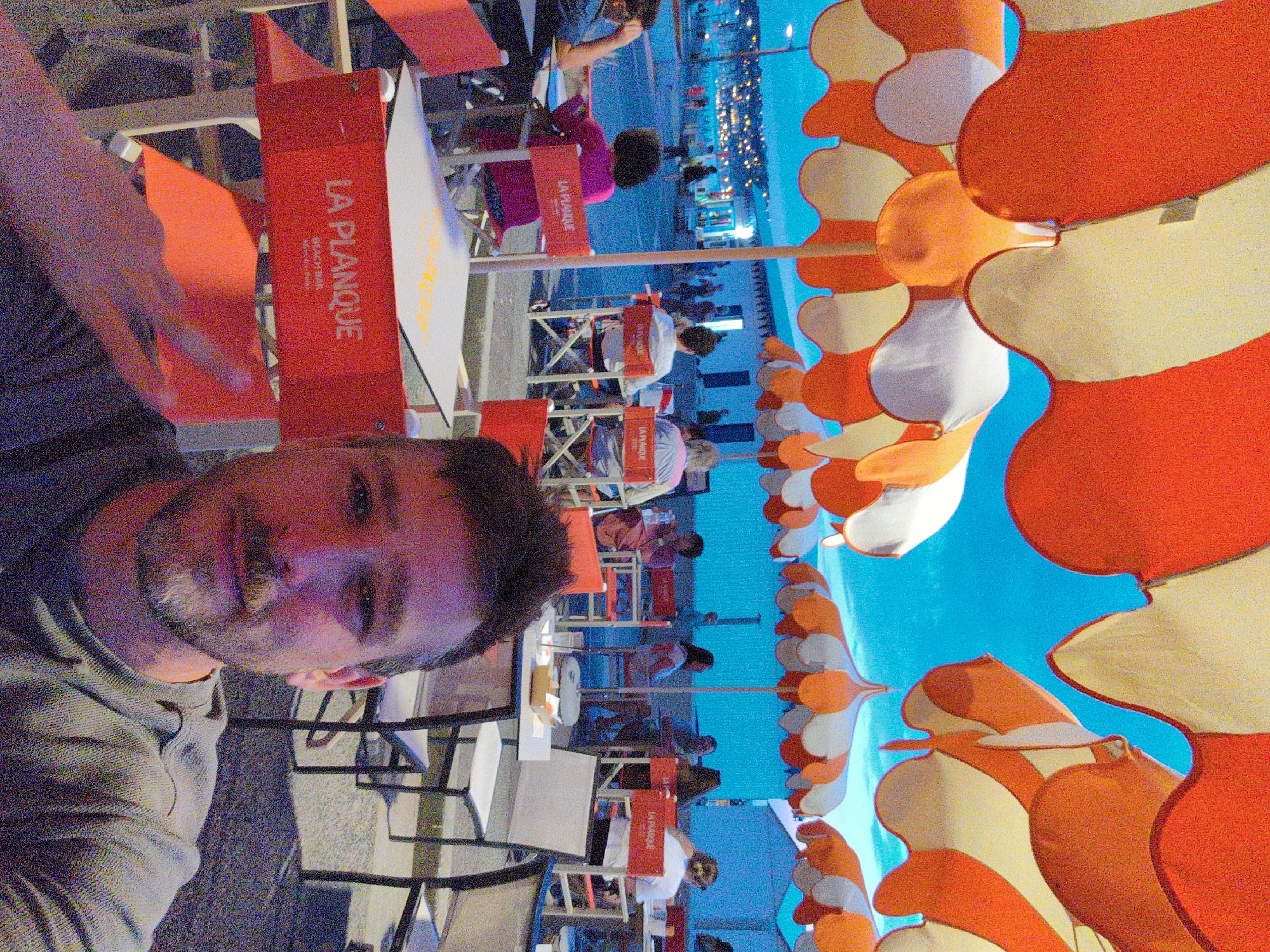 The image is a selfie taken at an outdoor cafe in Mers-les-Bains, France. The photographer, a man with a beard and mustache, is in the foreground, wearing a grey polo shirt. Behind him, several tables with red chairs are visible, some adorned with umbrellas. A sign on a red chair reads "LA PLANQUE BEACH BAR." In the background, people are walking along a waterfront promenade, with a town illuminated by lights stretching into the distance under a twilight sky. The cafe appears to be situated near a beach or harbor area.