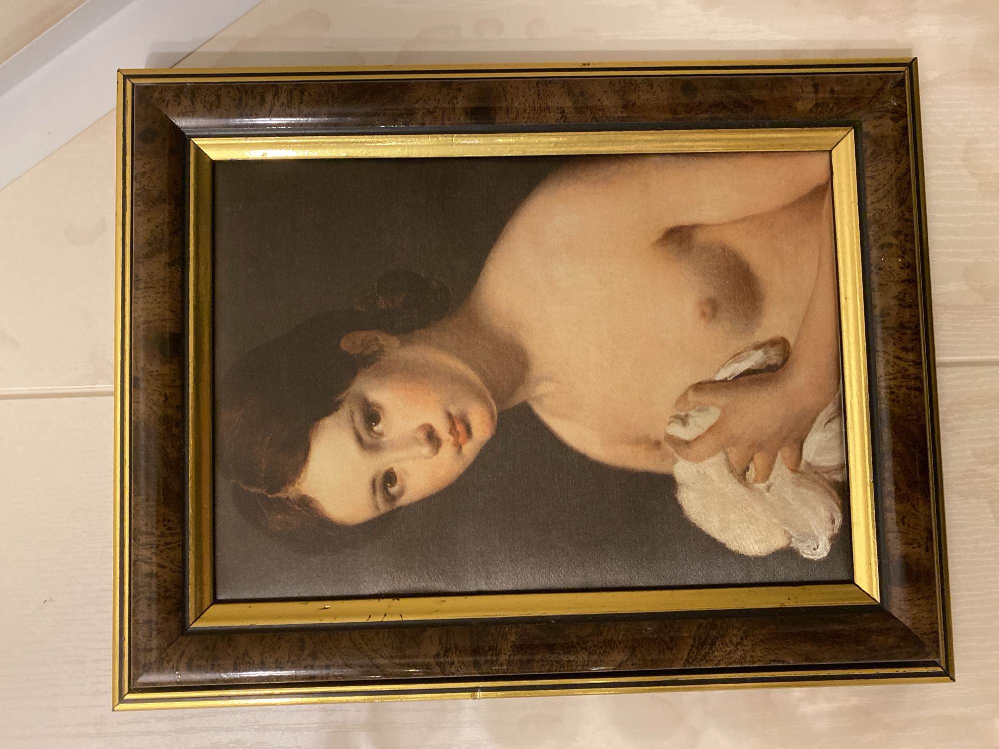 A vertically oriented framed portrait, depicting a partially nude young woman, hangs centered on a light-colored wall in the city of Unknown, Unknown. The painting features a young woman with dark brown hair parted down the middle and styled back, a fair complexion with rosy cheeks, and dark eyes. Her gaze is directed forward, with her head slightly tilted. She is depicted from the chest up, with bare shoulders and breasts, and is holding a substantial amount of white, possibly linen, fabric with both hands, positioned over her lower torso. The background of the painting is a dark, unadorned color.
The artwork is enclosed in a rectangular wooden frame. The frame consists of two primary sections: an outer, wider section of dark brown wood with a visible grain pattern, and an inner, narrower section of distressed gold. The gold trim exhibits multiple chips, scratches, and areas where the finish has worn away, revealing a darker underlayer. The dark brown section also shows minor scuffs.
The wall behind the framed artwork is light beige or cream-colored, displaying a subtle vertical texture. Several irregular, darker stains or marks are visible on the wall, particularly above the frame and to its right. A strip of white architectural trim or molding is partially visible along the upper right edge of the photograph.