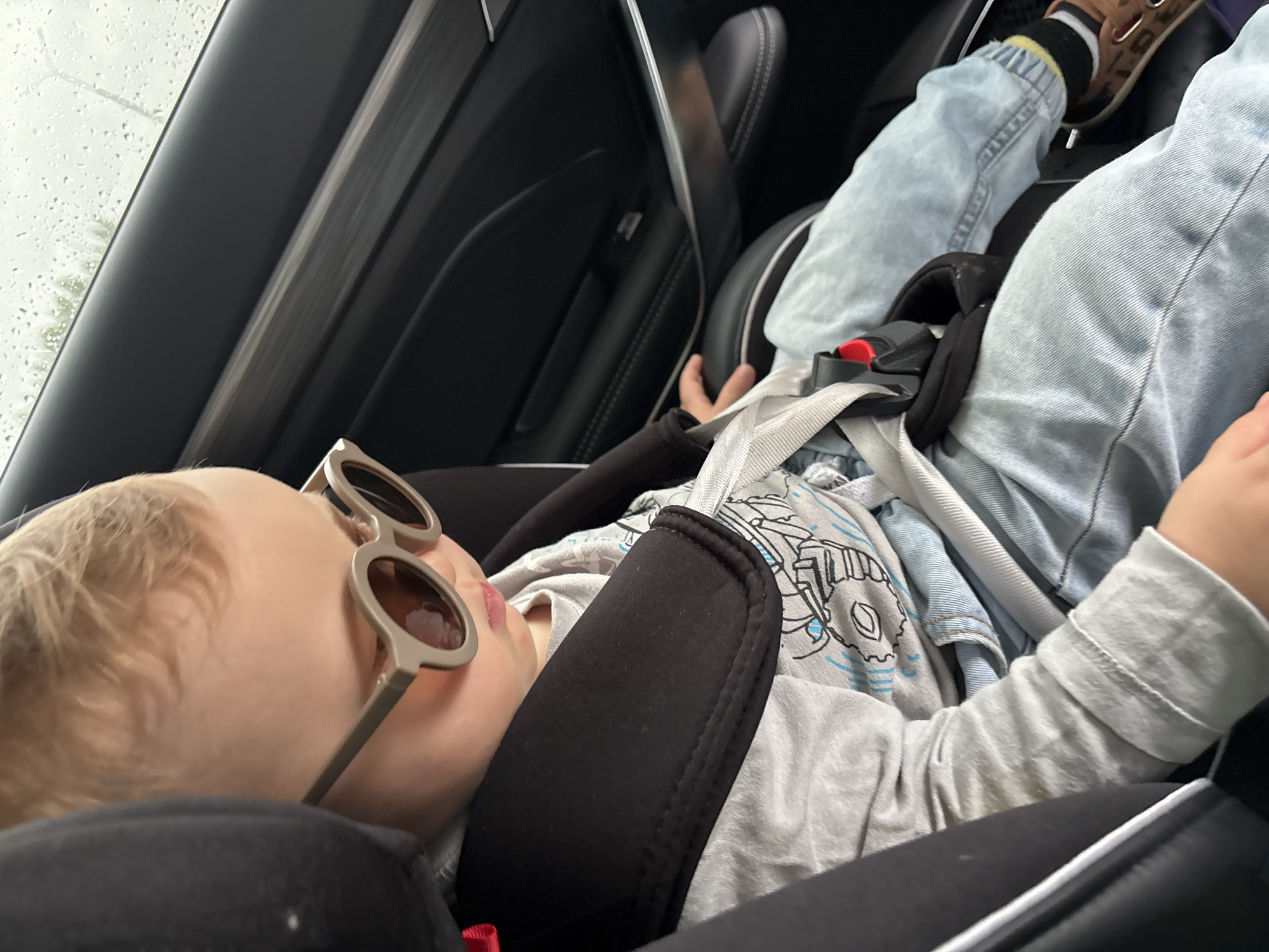 The image shows a young child, likely a toddler, seated in a car seat within a vehicle. The child is wearing a grey long-sleeved shirt with a graphic print, light blue jeans, sunglasses with a round, beige frame, and brown sandals with black socks. The child is looking downwards. The car interior is visible, including the black leather seat and door panel. Outside the window, raindrops are visible on the glass, indicating it is raining. The setting appears to be a street in the city of Naberezhnye Chelny, Russia.