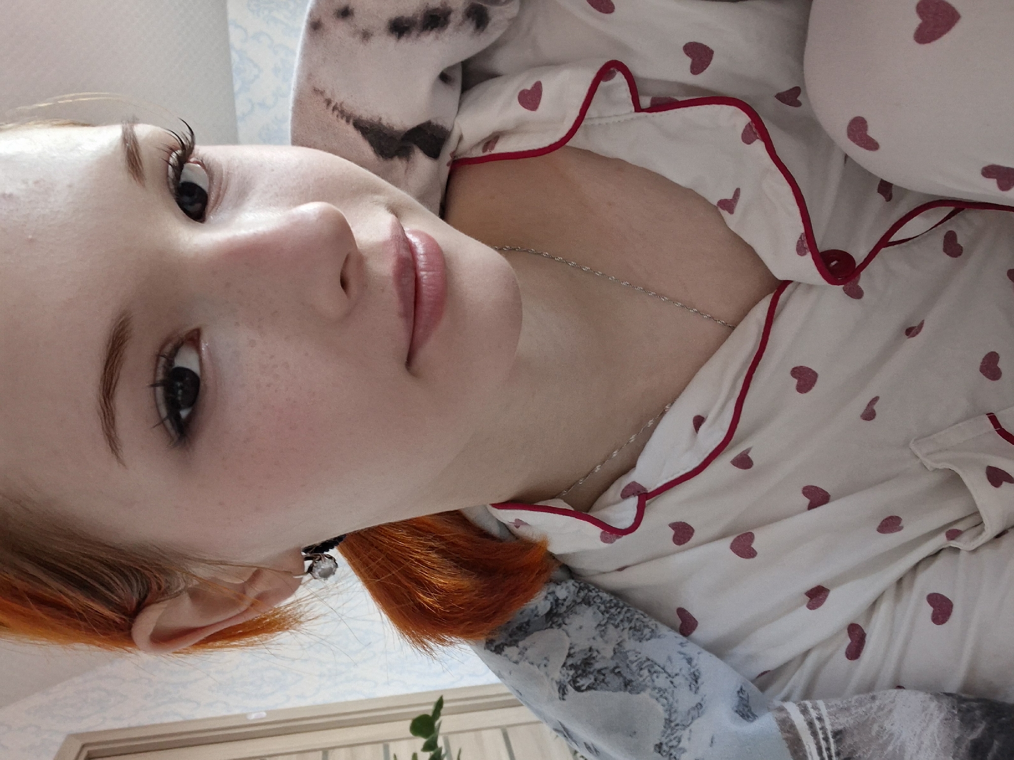 A selfie of a fair-skinned person with orange hair wearing an earring, a silver chain necklace, and a white pajama top with pink hearts and red trim, taken indoors. A pillow with a dalmatian print is visible behind the person's head. The location is indiscernible and cannot be identified as Chaikovsky, Russia, from the provided image.