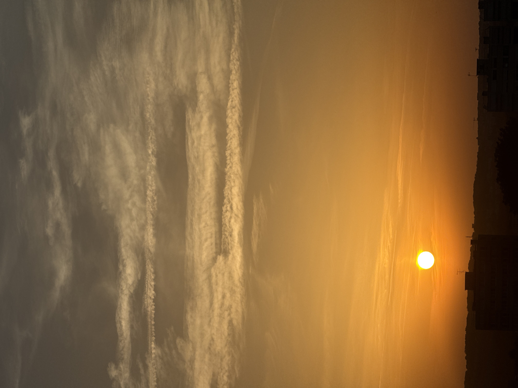 An aerial shot captures a sunset over an urban landscape. The sky is ablaze with hues of orange and yellow, transitioning to a darker grey near the top. Wispy clouds streak horizontally across the sky, some appearing almost like trails. The sun, a bright white orb, is low on the horizon, casting a warm glow. Below, the silhouettes of buildings and a dark, hilly terrain are visible against the vibrant sky. The image does not contain any people or discernible actions.