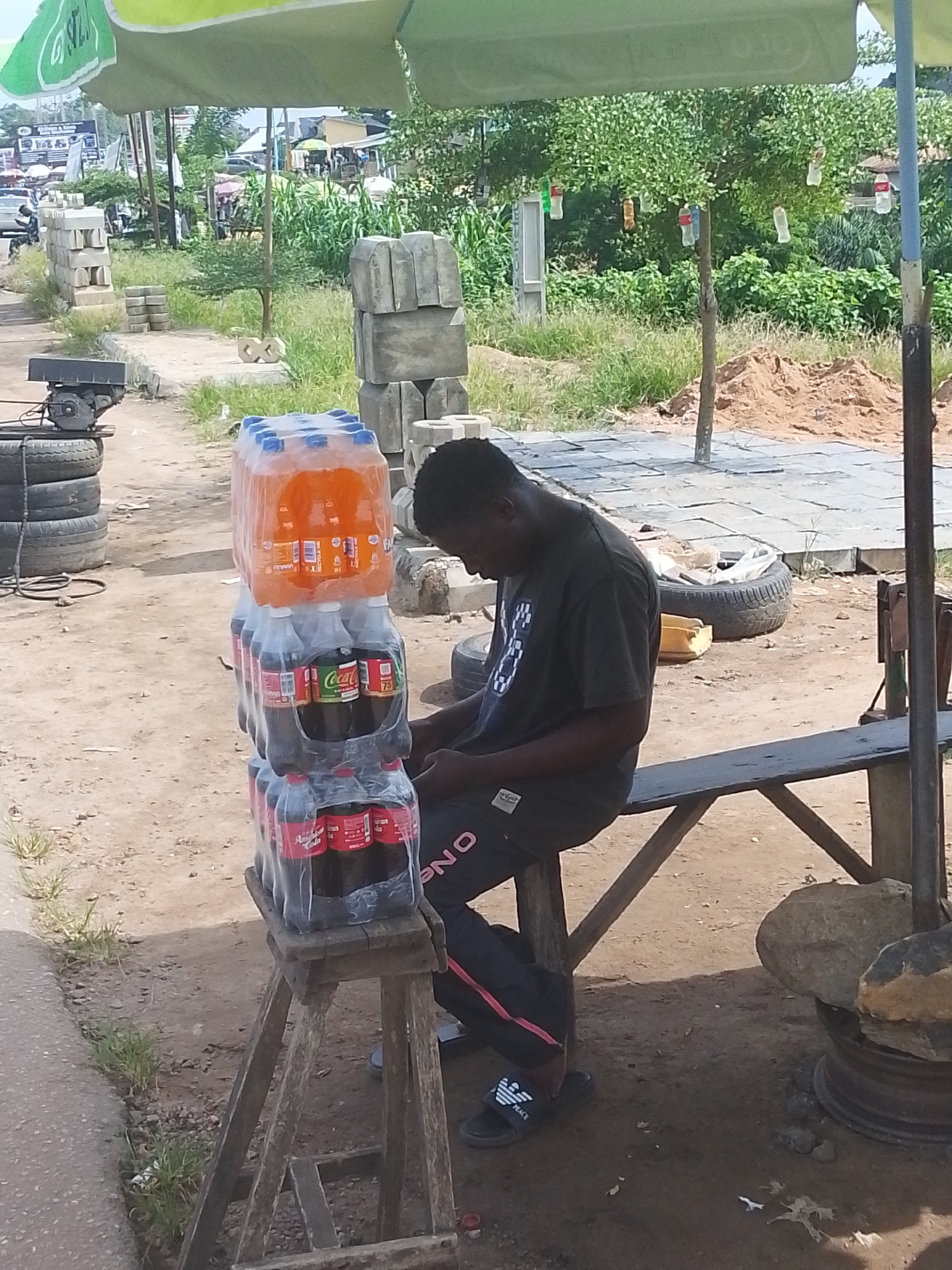 A young Black male wearing a dark t-shirt and dark pants with a red stripe down the side sits on a wooden bench under a green umbrella. He is looking down at his phone. To his left, on a wooden stool, are stacks of bottled drinks. The bottles are primarily Coca-Cola and a brightly colored orange soda. The ground is dirt and gravel, with some scattered debris. In the background, there are piles of cement blocks, some vegetation, and a distant road with vehicles and buildings. The scene appears to be an outdoor market or roadside stall in Abeere, Nigeria.