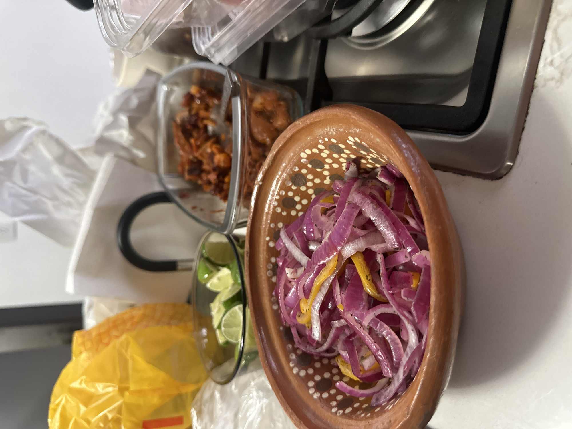 The image depicts a kitchen scene, likely in a home, in Naucalpan de Juárez, Mexico, evidenced by the typical Mexican pottery bowl. A brown ceramic bowl filled with sliced red onions and yellow peppers sits prominently in the frame. To the left are two glass containers, one with sliced limes and the other holding a reddish-brown food item that could be chili or a similar ingredient. Behind these is a partially visible container with a clear lid, suggesting it contains more food ingredients. A yellow bag and crumpled white plastic bag are at the bottom left. In the upper right, a stainless-steel stove is partially visible. The scene is set on a white countertop.
