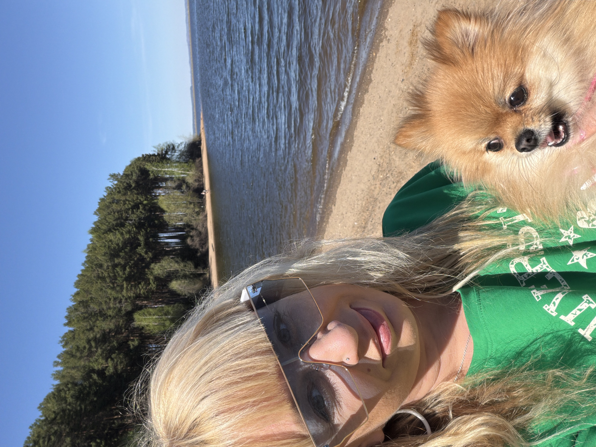A rotated eye-level shot presents a fair-skinned woman with blonde hair wearing sunglasses and a green t-shirt, holding a small brown Pomeranian dog with its mouth open. Behind them is a body of water bordered by a sandy beach and a line of trees under a clear blue sky. The scene is likely set in Chaikovsky, Russia, as requested.