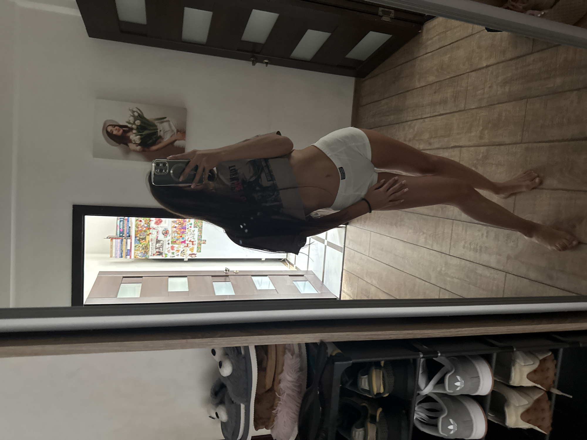 A full-body, mirrored selfie shows a young woman standing in what appears to be a residential hallway. She is wearing a cropped t-shirt and white boxer shorts. To the left of the frame, a multi-tiered shoe rack is visible, containing several pairs of shoes and slippers. In the background, a door with glass panels is slightly ajar, revealing a glimpse of another room. On the wall to the right of the woman, a framed picture of a woman holding flowers is displayed. The flooring is made of light-colored wood-effect tiles. The overall environment suggests an indoor domestic setting. There is no specific information in the image to indicate the location is in Chișinău, Moldova.