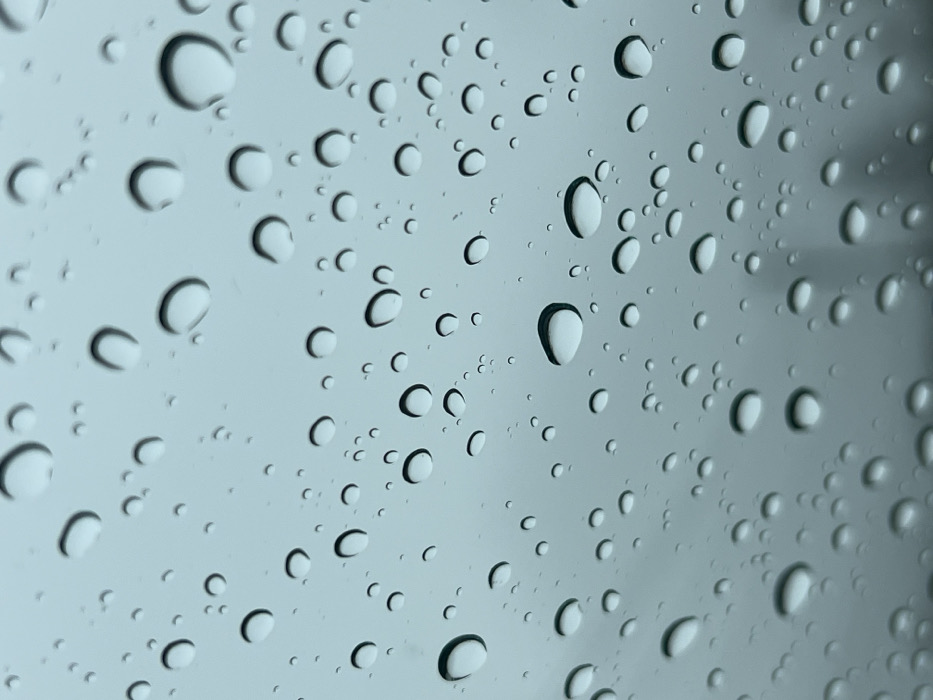 A close-up shot depicts numerous raindrops clinging to a smooth, translucent surface, possibly glass. The drops vary in size, from small to medium, and are uniformly distributed across the frame. Each droplet features a crescent-shaped highlight, indicating a light source positioned above. The background appears blurred and indistinct, contributing to the focus on the water droplets. The color palette is dominated by shades of gray and white, suggesting overcast or indoor lighting conditions.