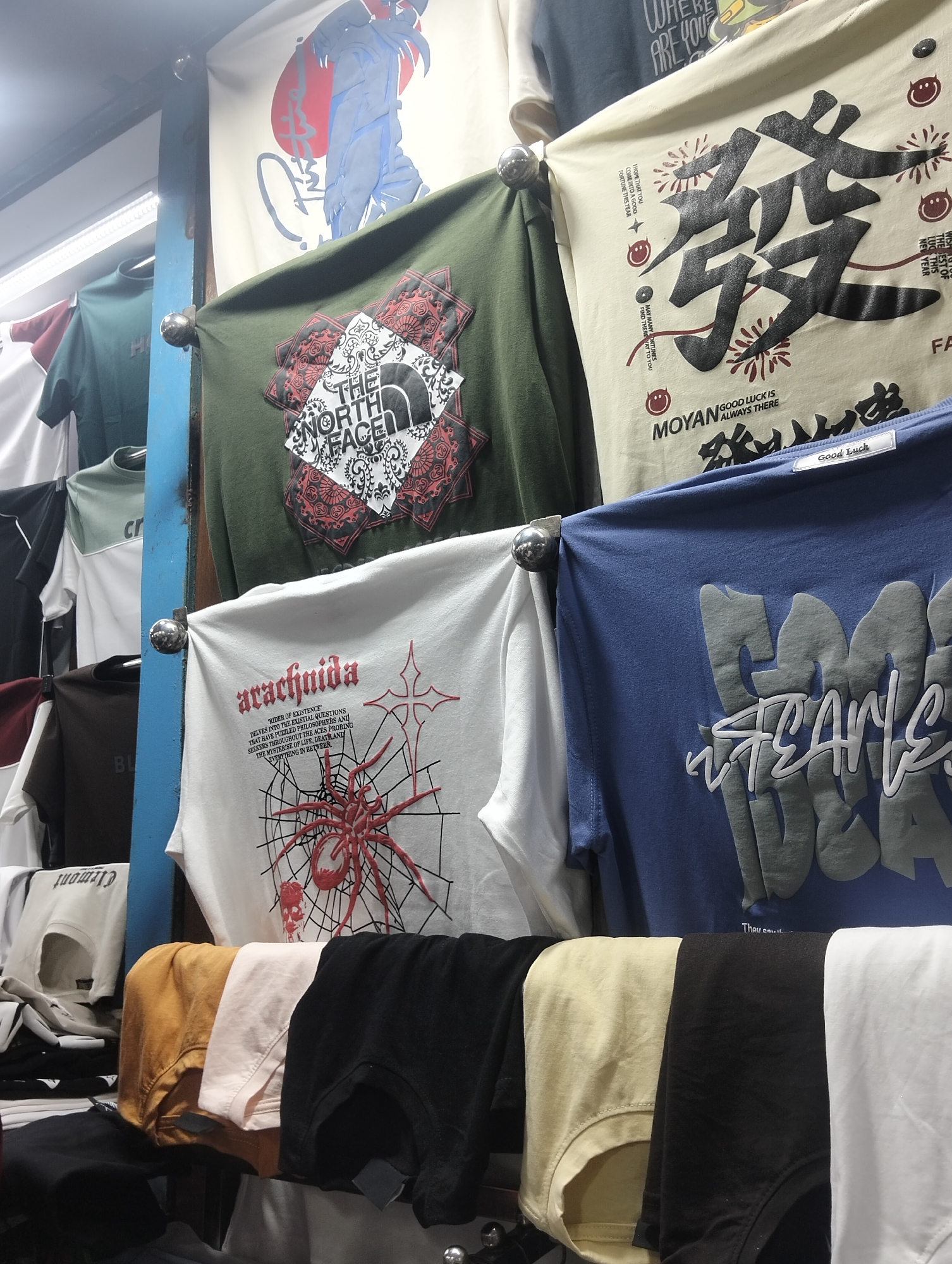 The image shows a rack of t-shirts for sale in a shop. The t-shirts have various designs, including "The North Face", a spider, Chinese characters, and graffiti-style text. The rack has shirts in a range of colors. The location is Dhaka, Bangladesh.