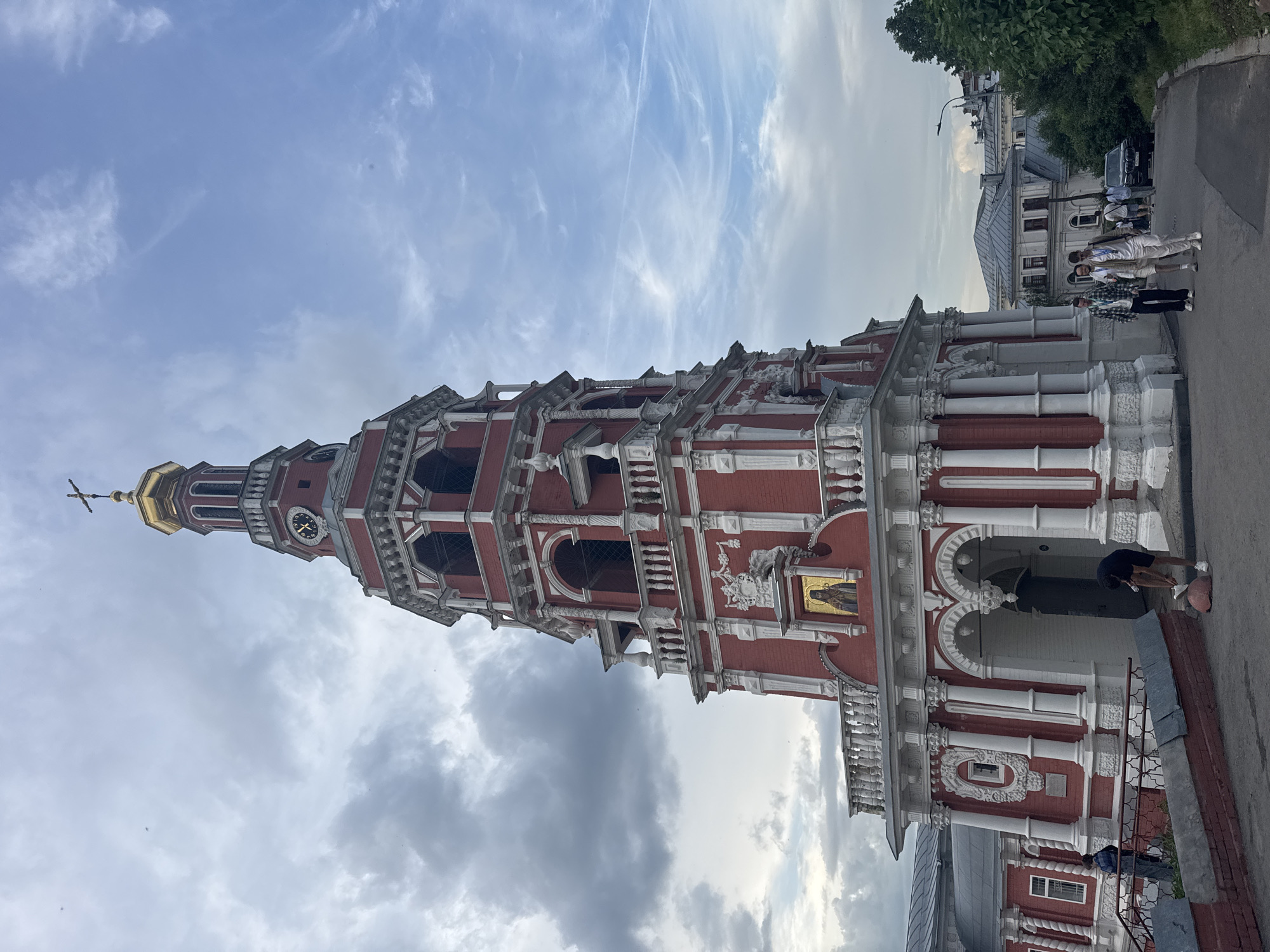 The image shows the Church of the Nativity of John the Precursor in Nizhny Novgorod, Russia. The church is a tall, red and white building with a golden spire. The sky is partly cloudy. There are a few people walking on the sidewalk next to the church. One person seems to be retrieving a ball near the bottom of the church.