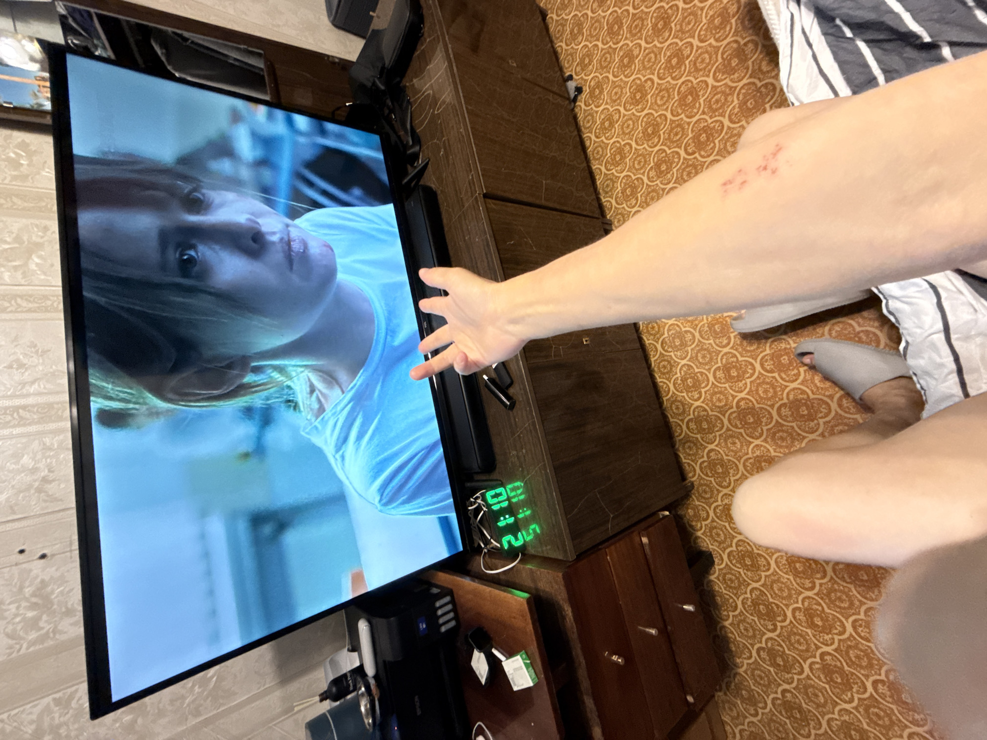 An indoor, eye-level, medium shot captures a person's right arm and leg in the foreground, positioned on a patterned carpet and partially on a striped duvet. The person's right arm is extended towards a television screen showing a close-up of a woman's face. A digital clock on a stand below the TV displays "21:10". To the left of the clock is a printer and a wooden drawer unit. The scene appears to be a living room. The image provides no information about the city of Khimki, Russia.