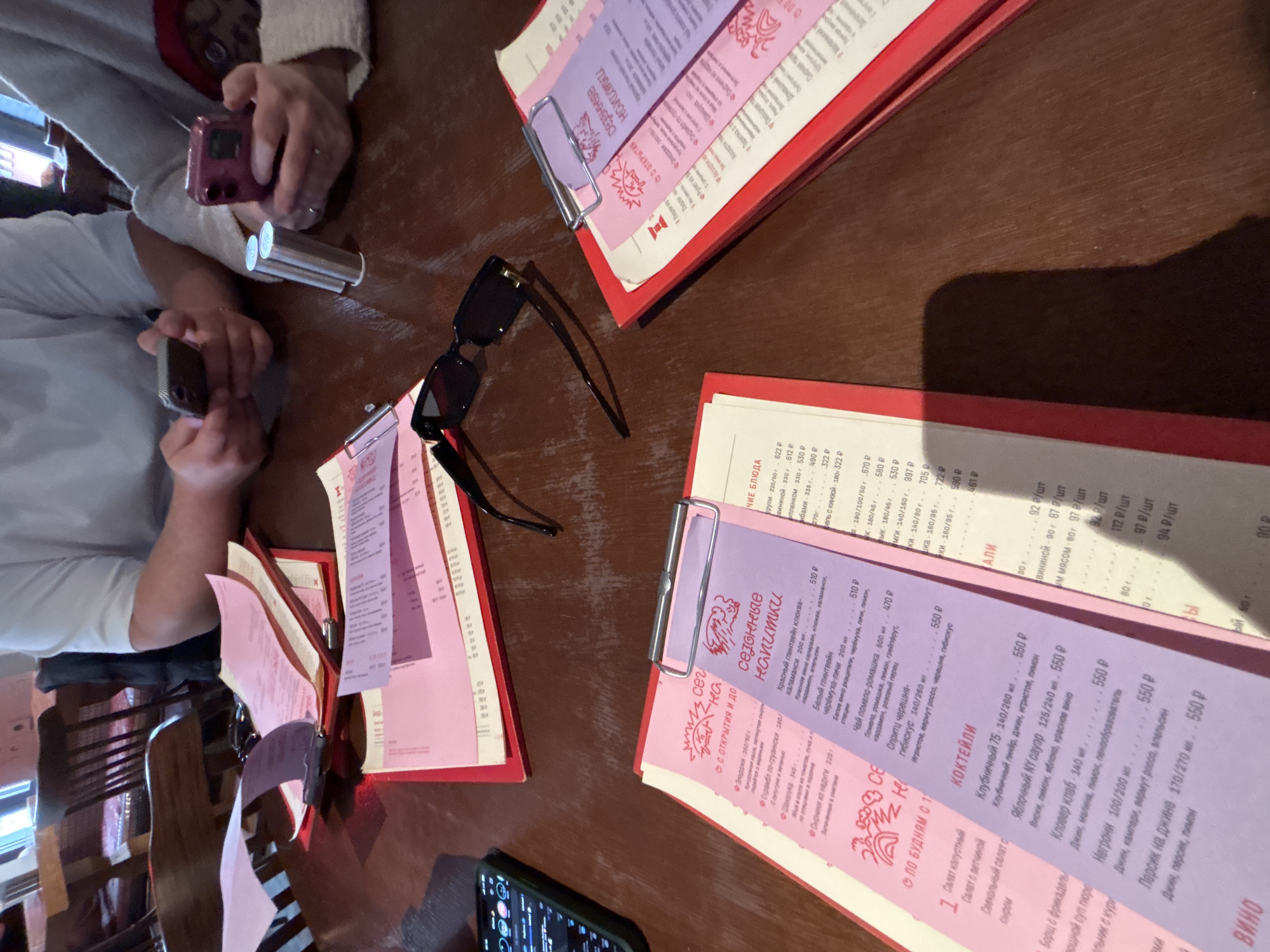 The image shows a restaurant table with menus, sunglasses, phones, and hands of two people, likely in Moscow, Russia. The menus are red-bordered and contain Russian text and prices in rubles. The table appears to be wooden and is cluttered with items. One person is using a pink smartphone, while the other has a black smartphone. There is also a cylindrical object on the table, possibly a vaporizer or lighter.