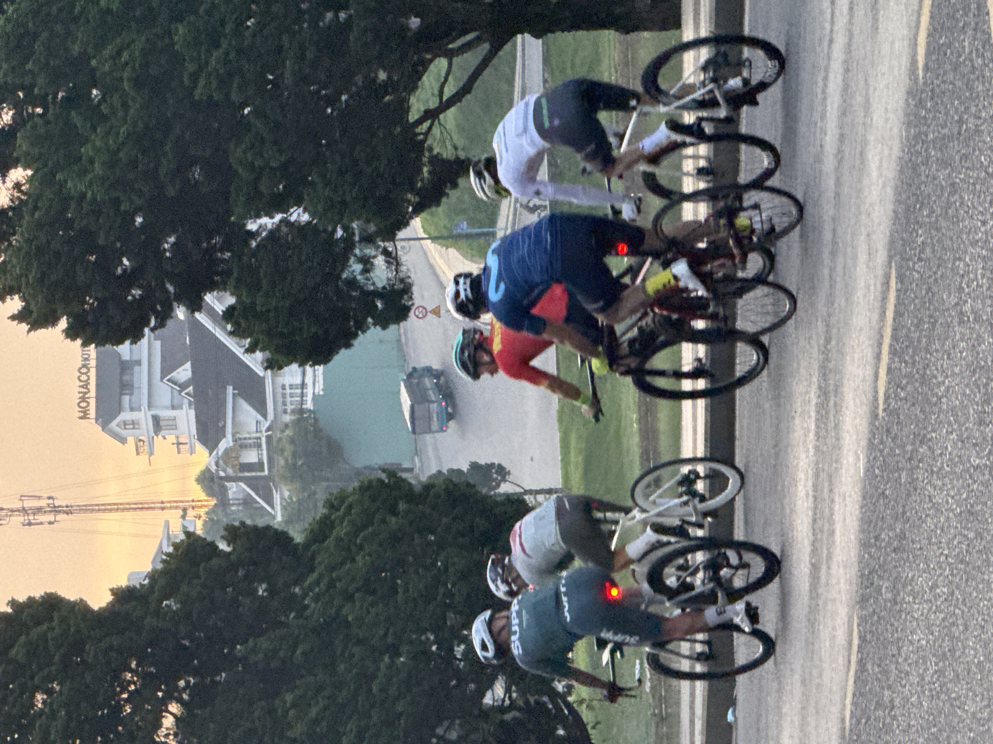 A group of cyclists are riding in a road race. The cyclists are wearing helmets and cycling gear. The scene is set on a paved road with a yellow line down the middle. There are trees on the left side of the road, and buildings in the background. The time of day appears to be late afternoon or early evening, as indicated by the warm, soft lighting. The weather appears to be clear. The word "MONACO" is visible on a building in the background. One cyclist has a red light on the back of their bike. The cyclists are positioned in a staggered formation, suggesting they are competing. The overall impression is one of athletic activity in a scenic, possibly urban or suburban, setting.