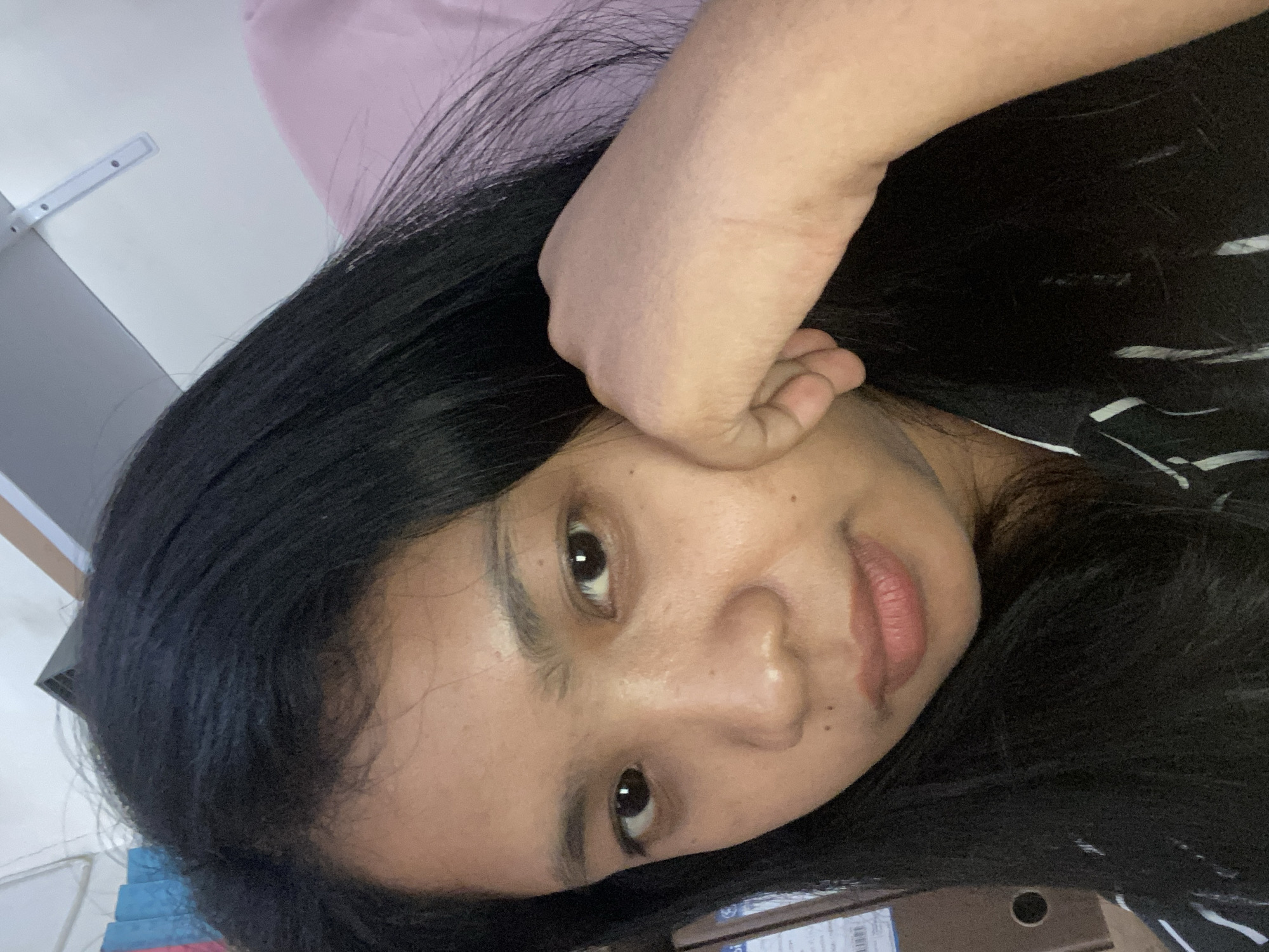 A young woman with long, black hair rests her face on her hand. She has brown eyes, and her skin tone is medium. She is wearing a black and white striped shirt. The location is Negara, Indonesia, indicated by the presence of a shipping box with associated labels commonly found in Indonesia, but not explicitly visible. Other objects in the scene include a pink surface in the background and stacked items on a surface. The image is taken from a close-up, slightly tilted perspective.