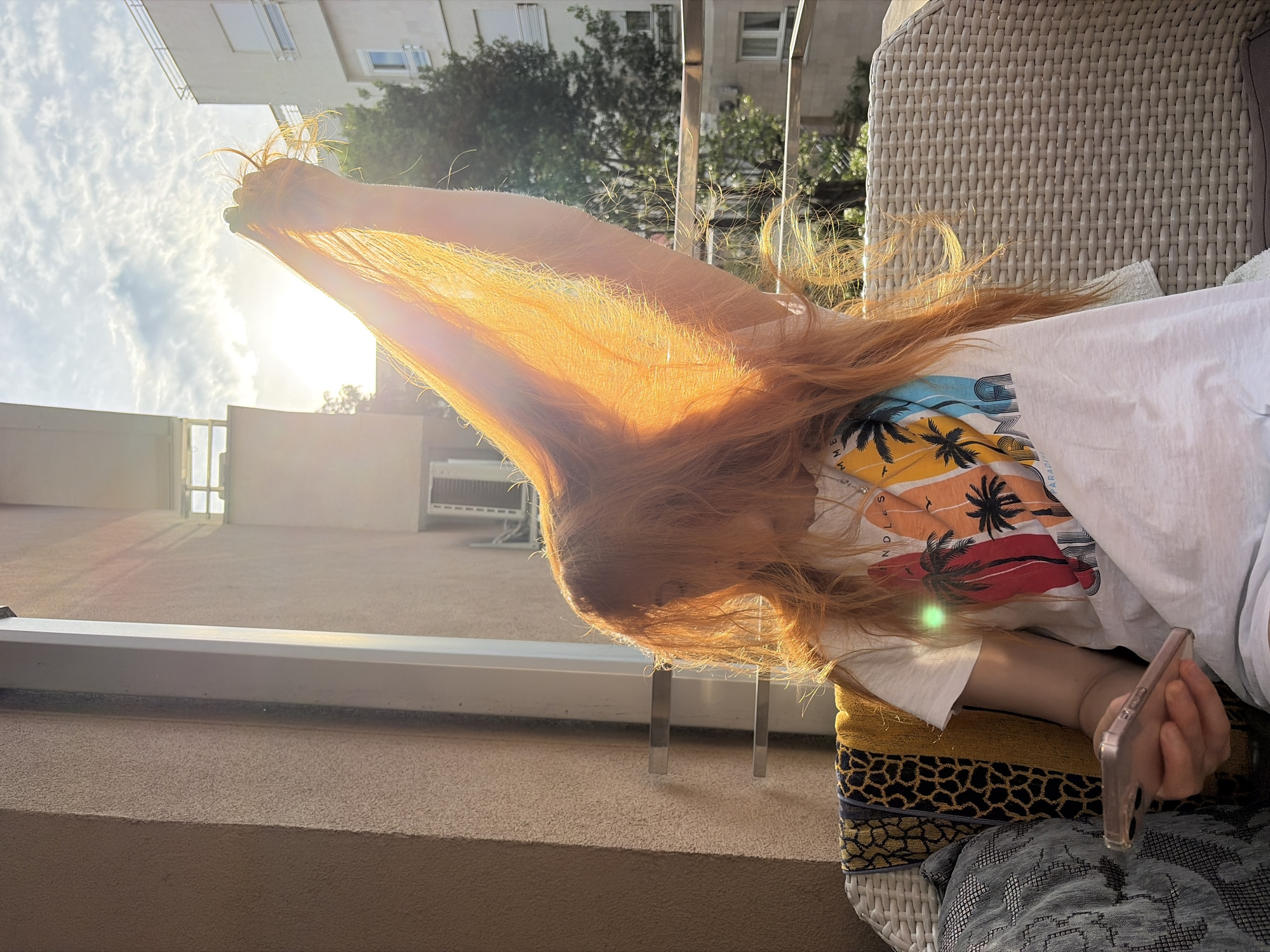 A young person with long, auburn hair is sitting on a wicker chair on a balcony in what appears to be Budva, Montenegro. The hair is held up in front of their face, illuminated by the setting sun, creating a bright orange glow. They are wearing a white t-shirt with a colorful palm tree design and are holding a smartphone in their hand. The balcony is made of beige concrete, and there is a metallic railing visible. In the background, there is a building and some trees. The sky is partly cloudy with a bright sun.