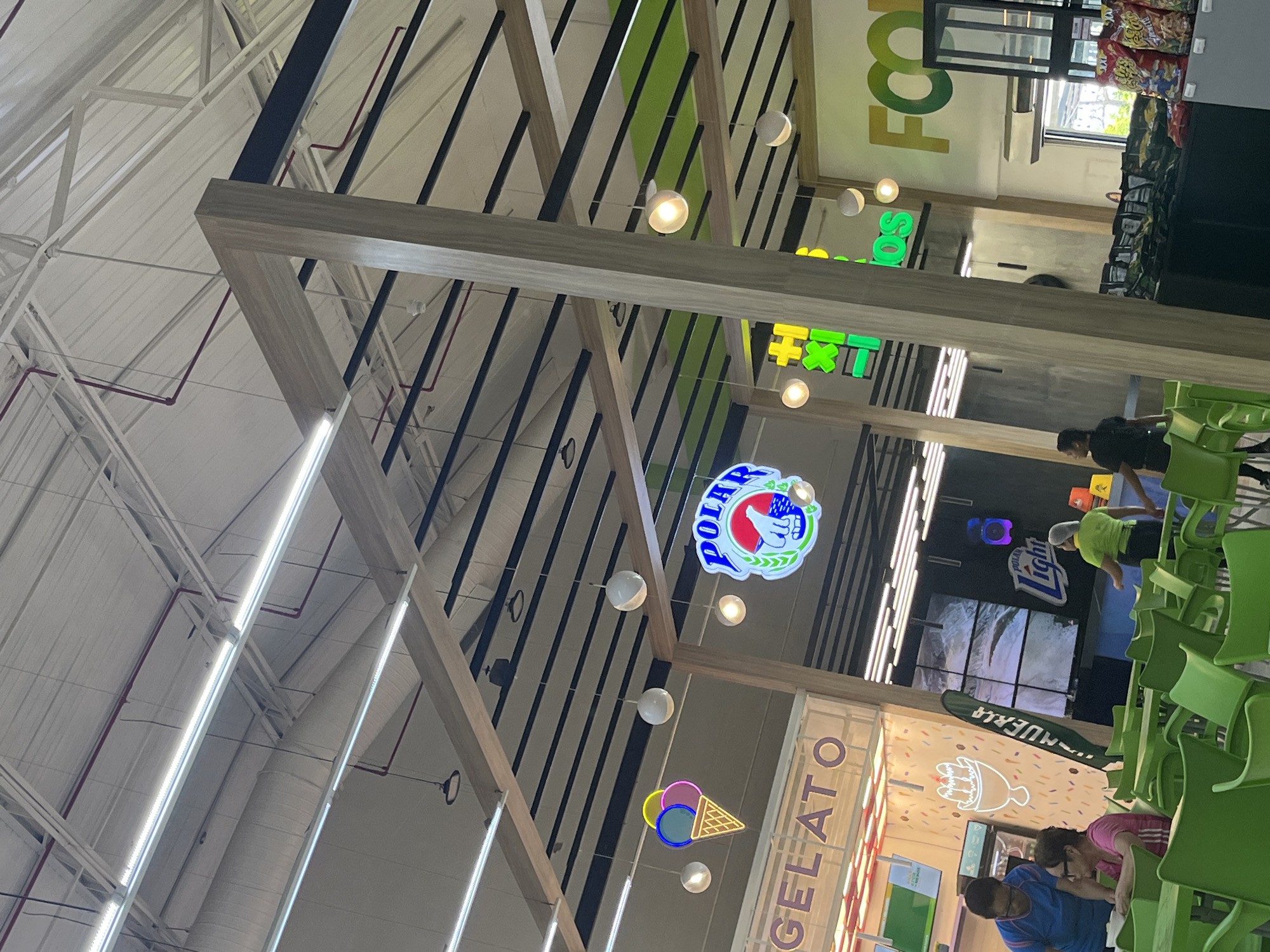 The image depicts the interior of a supermarket or food court in Porlamar, Venezuela. Overhead, a wooden framework with black supports spans the space, bearing signs for "FOOD," "Polar," and "GELATO." Spherical light fixtures are suspended from the structure.
To the right, there are green plastic chairs arranged near food stalls or vendors. Individuals are seen in the vicinity: one wearing a lime green shirt, another in a black shirt, and two individuals dressed in pink and blue near the "GELATO" sign. Shelves stocked with consumer goods are partially visible in the background.