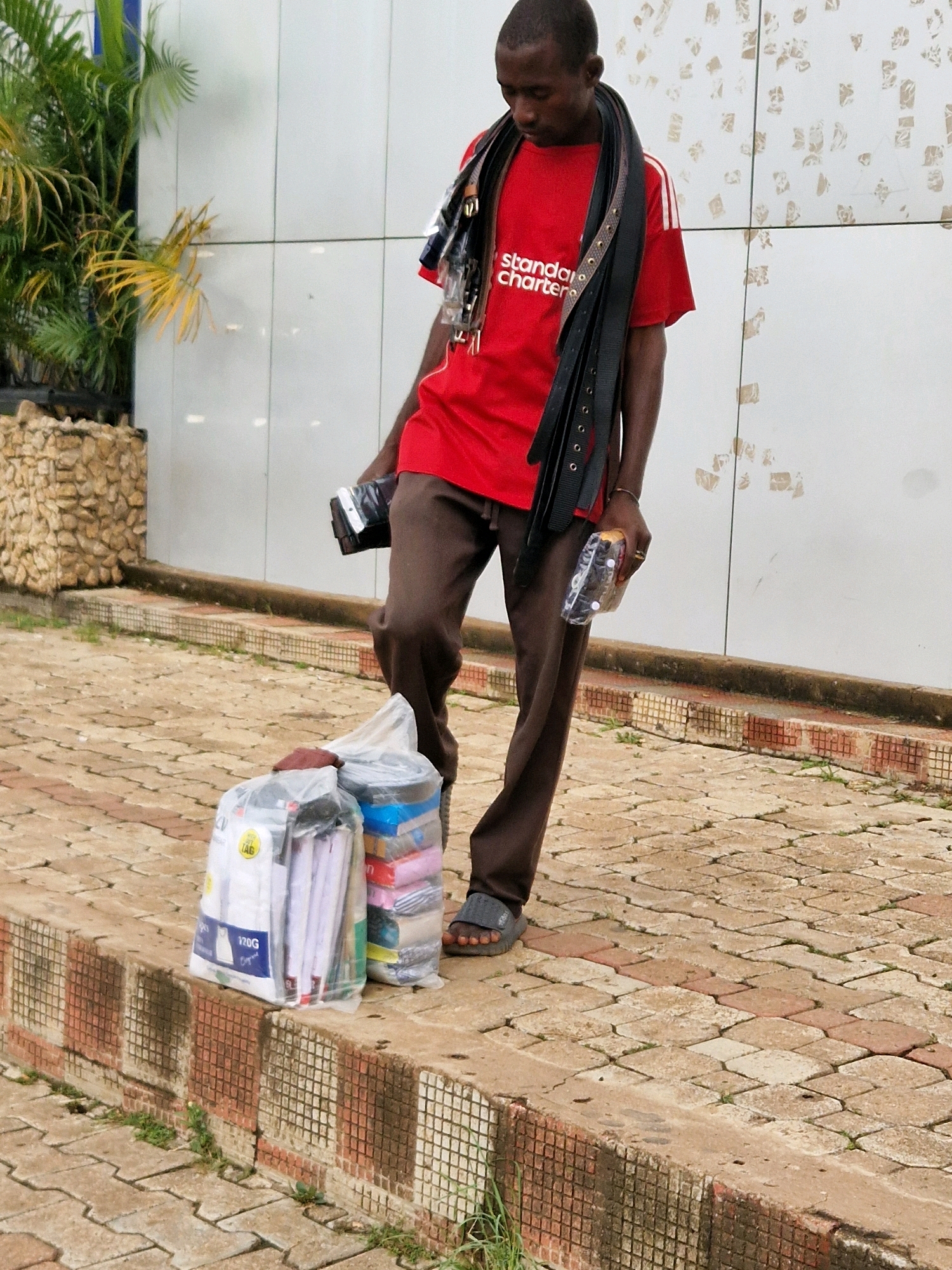 A young Black male stands on a paved sidewalk in Jos, Nigeria, looking down. He is wearing a red t-shirt with "Standard Chartered" printed on it and brown trousers. A black leather belt is draped around his neck and shoulders, with several other belts hanging from it. He is holding a black wallet or small bag in his right hand and a plastic-wrapped item in his left hand. In front of him, on the edge of the sidewalk, are two large plastic bags containing various items. One bag appears to hold stacks of paper, possibly notebooks or reams of paper, and the other contains what looks like folded clothing or textiles. The background shows a white wall with a textured pattern and some greenery from a plant to the left. The ground is paved with interlocking bricks.