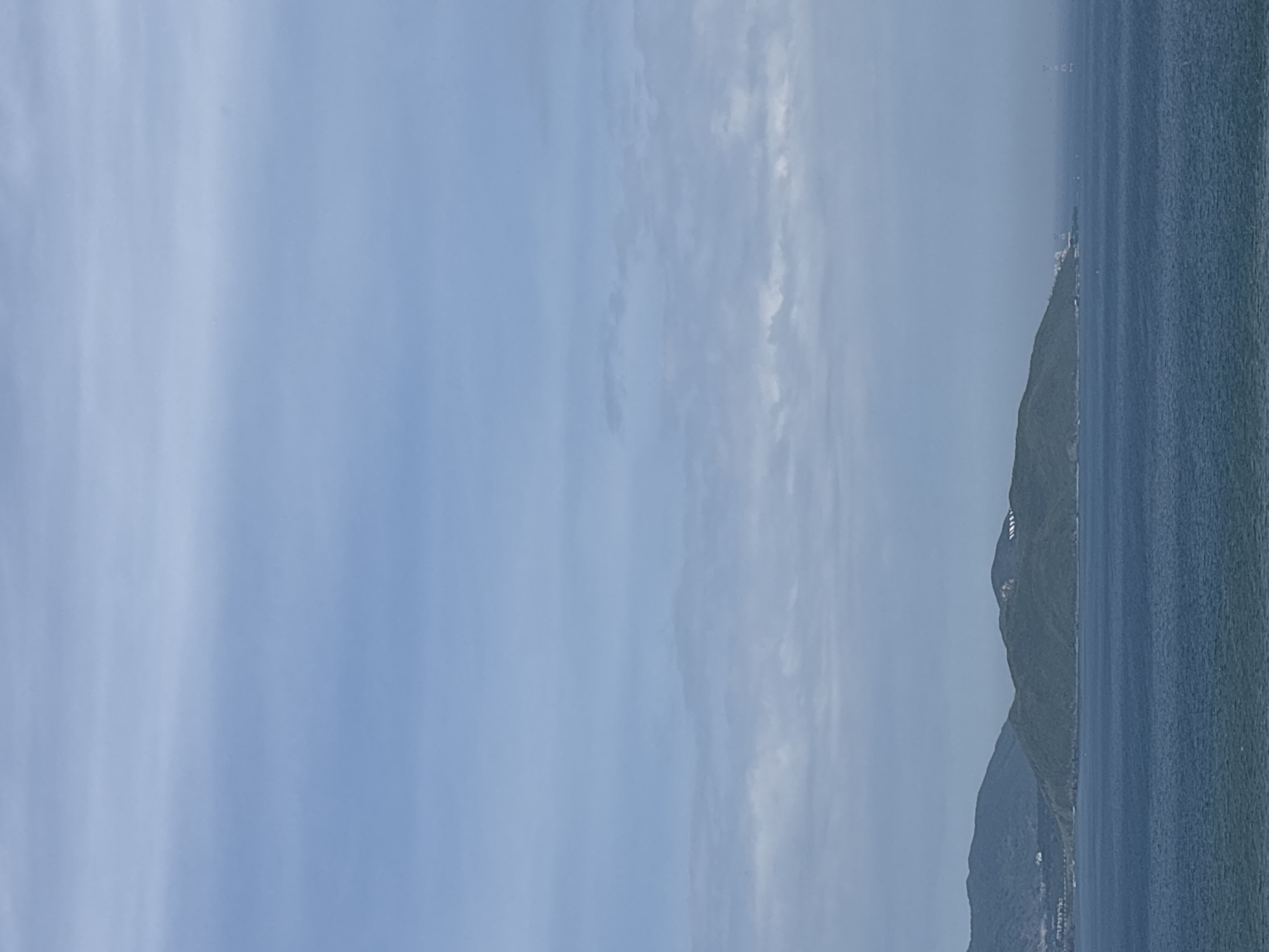 The image shows a wide shot of a coastal landscape under a cloudy sky. In the foreground, a deep blue body of water stretches across the frame, with visible waves. In the middle ground, a chain of green, hilly islands runs parallel to the coast. The largest island in the center has structures on its peak, including what appears to be white lettering that spells out "VINPEARL." Further to the right, a distant pylon or tower structure is visible on the horizon. The background is dominated by a pale blue sky filled with streaks of white and gray clouds. The overall impression is a serene, natural scene with hints of human development.
