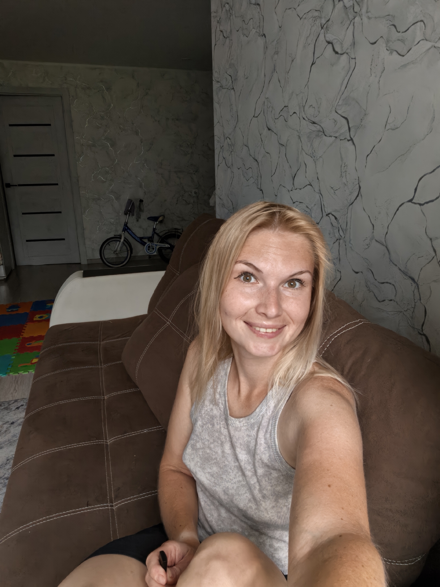 A blonde woman in her 20s sits on a brown couch in a room. She is wearing a gray tank top and black shorts. The woman is smiling and looking at the camera. The background of the image shows a wall with a marble-like pattern, a door, and a child's bicycle. There are also colorful puzzle mats on the floor. The scene appears to be indoors in a residential setting. The location of Sterlitamak, Russia is not ascertainable from the provided image.