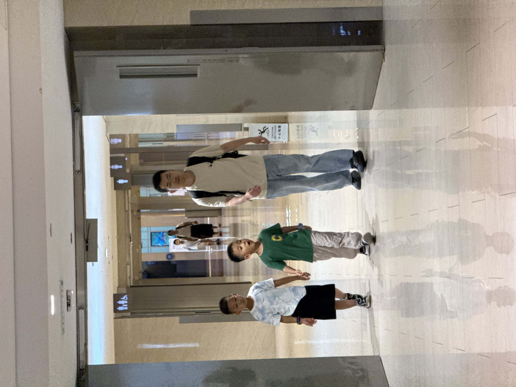The image depicts an indoor hallway with polished tile flooring that reflects the overhead lighting. On the left, two young boys walk hand-in-hand. The boy on the left wears a blue and white tie-dye t-shirt and a black skirt. The boy on the right wears a green t-shirt and light-colored pants. Behind them, a man with glasses wearing a tan t-shirt and blue jeans, carrying a black backpack, walks forward. In the background, further down the hallway, there are restroom signs and several other individuals walking. A yellow and white caution sign indicating a wet floor is visible near the man. The architecture suggests a modern public building, potentially a shopping mall or transit station. The scene appears to be in the city of Yuzhong District, China.