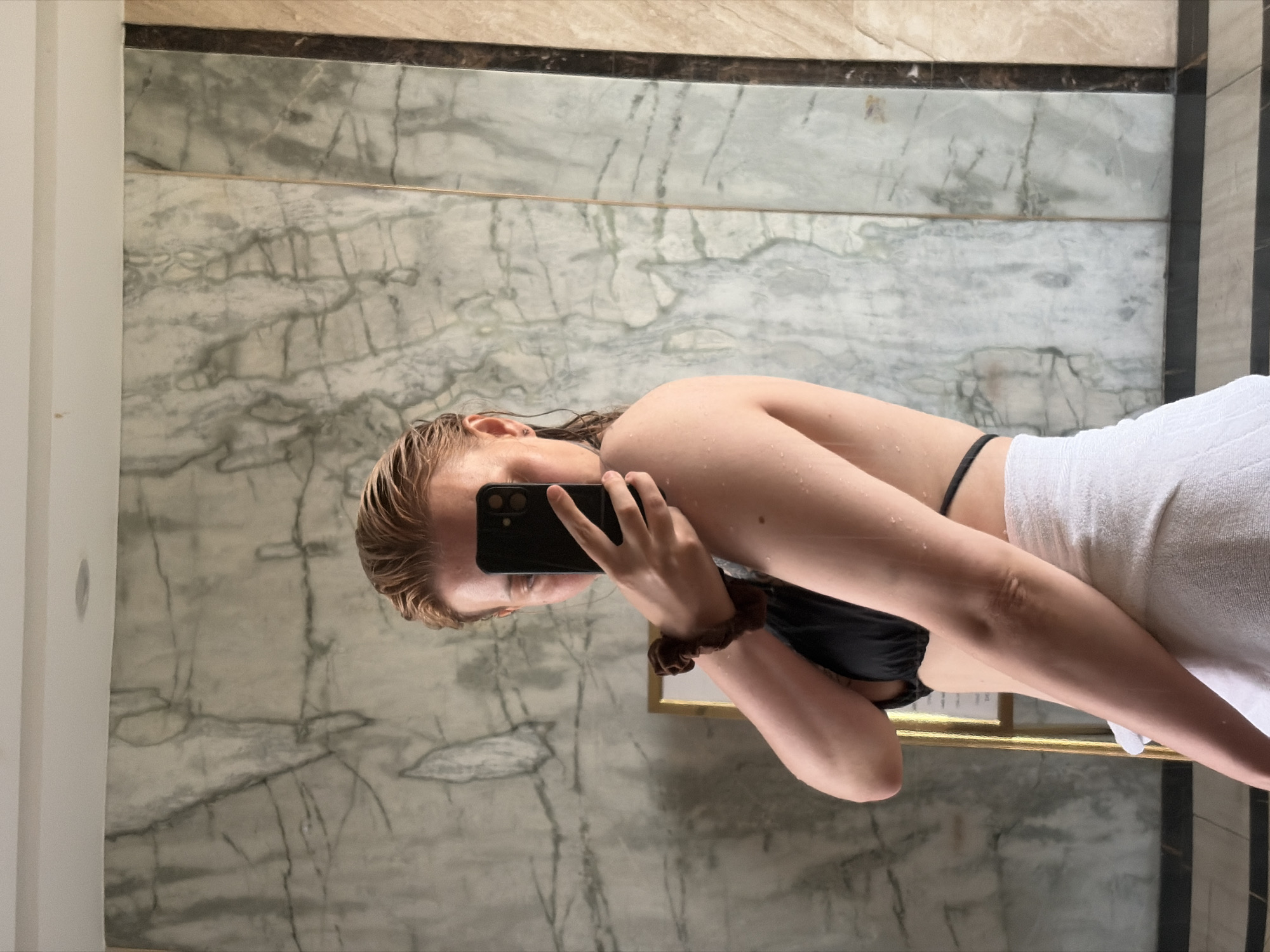The image, presented in a landscape orientation but depicting an upright subject, shows a mirror selfie taken by a person, likely a young woman. Her head is positioned towards the upper left, and her body extends diagonally across the frame.
The individual has wet, light-colored hair (blonde or light brown) that appears slicked back, with visible water droplets on her hair, shoulders, and arms, suggesting she has recently showered or been in water. She is wearing a black bikini top and has a white, textured towel wrapped around her lower body. A brown scrunchie is visible on her right wrist. Her face is mostly obscured by a black smartphone, which she holds up to capture the reflection. The phone has visible camera lenses on its back.
The background consists primarily of elegant light-colored marble walls with prominent gray veining. A thin golden horizontal trim runs across the wall behind the person's waist. Above this, there's a narrow dark brown horizontal strip, topped by a wider, lighter beige or tan section, likely also marble or tile. To the far left, a vertical white partition or pillar is visible, while a dark vertical stripe on the far right suggests part of a door frame or wall panel. The setting appears to be an indoor space, possibly a luxurious bathroom, changing room, or spa area, consistent with a resort or hotel in Đà Nẵng, Vietnam. There is no visible text in the image.