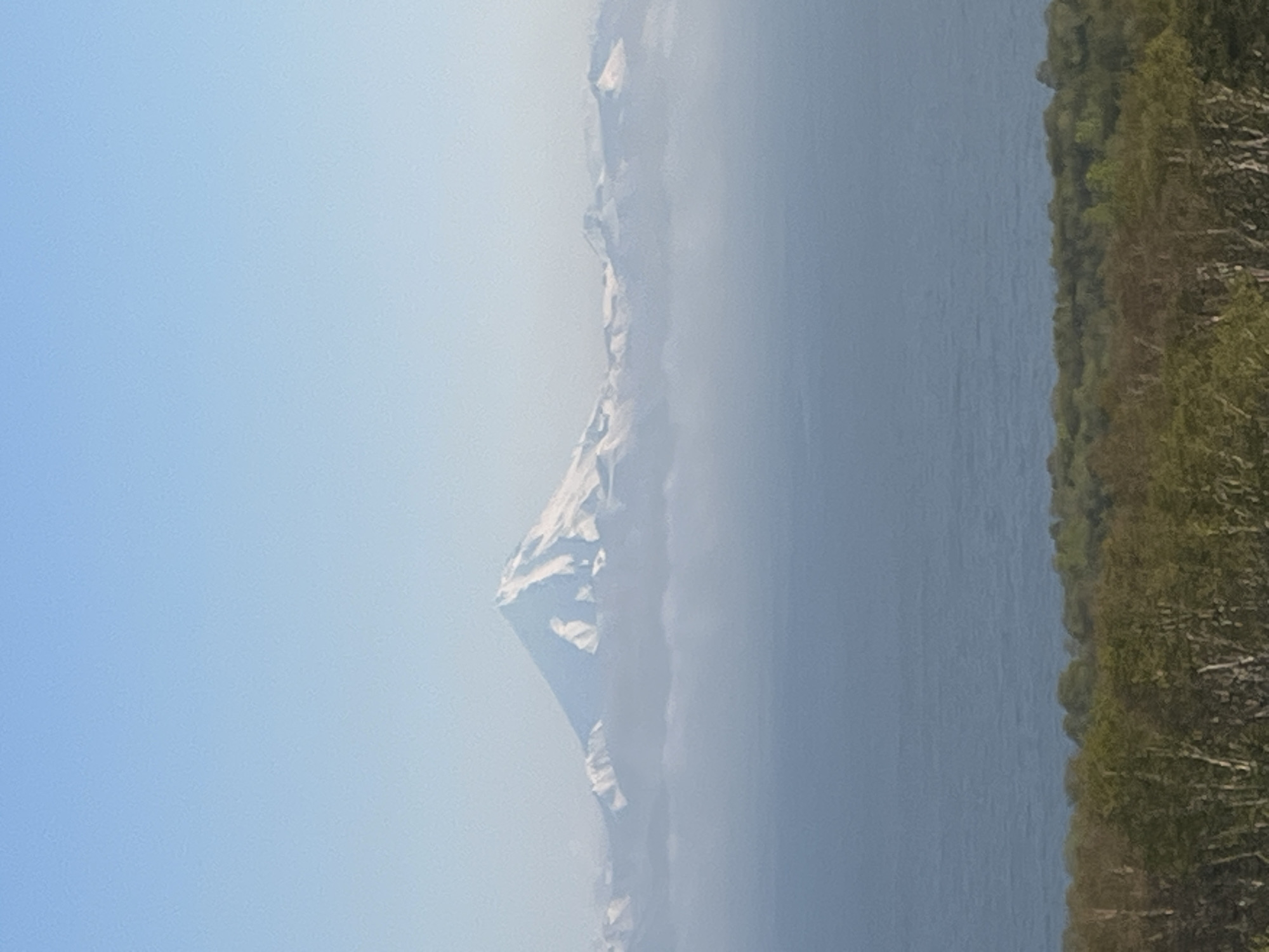 The image shows a majestic, snow-capped mountain peak rising above a layer of clouds, with a vast body of calm, blue water in the foreground. Below the water, a dense, green forest stretches across the landscape. The sky above is a clear, soft blue, with a gradient that lightens towards the horizon. The mountain is the central focus, its white summit stark against the pale blue sky, suggesting a cold, high-altitude environment. The clouds form a soft, ethereal barrier between the mountain and the water, creating a sense of depth and distance. The forest in the foreground provides a grounding element, its varied shades of green and brown indicating a temperate climate. The overall scene evokes a sense of natural beauty and tranquility. While the image does not contain any people or specific man-made structures, the presence of the ocean and forest suggests a coastal or mountainous region. The description of the location in the city of Крутобереговый, Russia, if accurate, would place this scene in a specific geographical context, implying it is a natural landscape within or near that city, potentially a view from a higher elevation overlooking the coast and the surrounding wilderness.