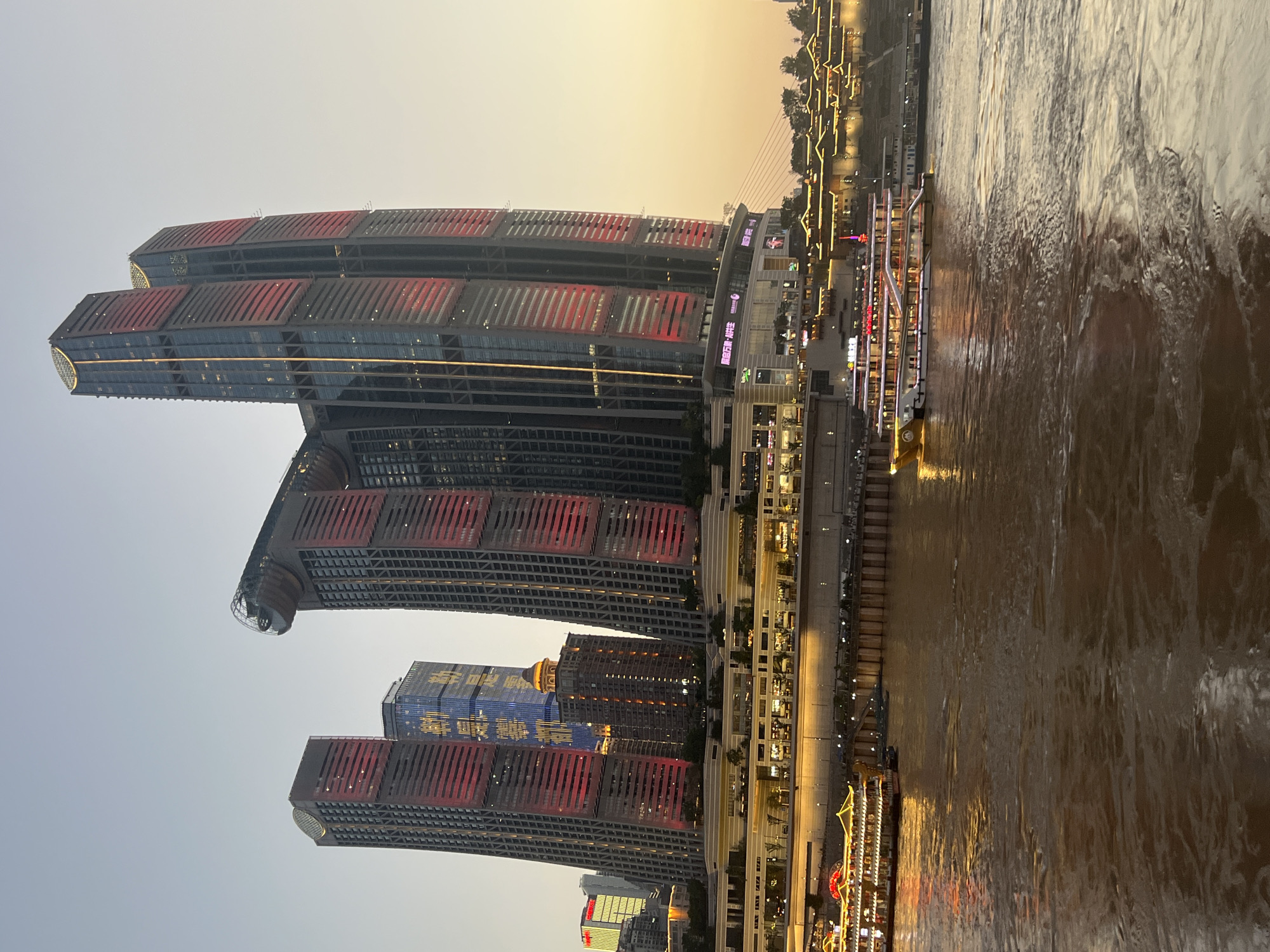 This is a photograph taken at dusk from a boat on a wide, brown river. In the background, the Nan'an District of Chongqing, China, is visible. Dominating the skyline are several modern skyscrapers, some of which are illuminated with red and yellow lights, creating a striking contrast against the pale sky. One building features a distinctive circular structure near its top. A waterfront promenade with illuminated buildings and traditional architecture is visible along the riverbank. Two large passenger boats are on the water, one in the foreground and another further back, both lit up and reflecting on the murky river surface. The sky is a soft gradient of yellow and light blue, indicating either sunrise or sunset.