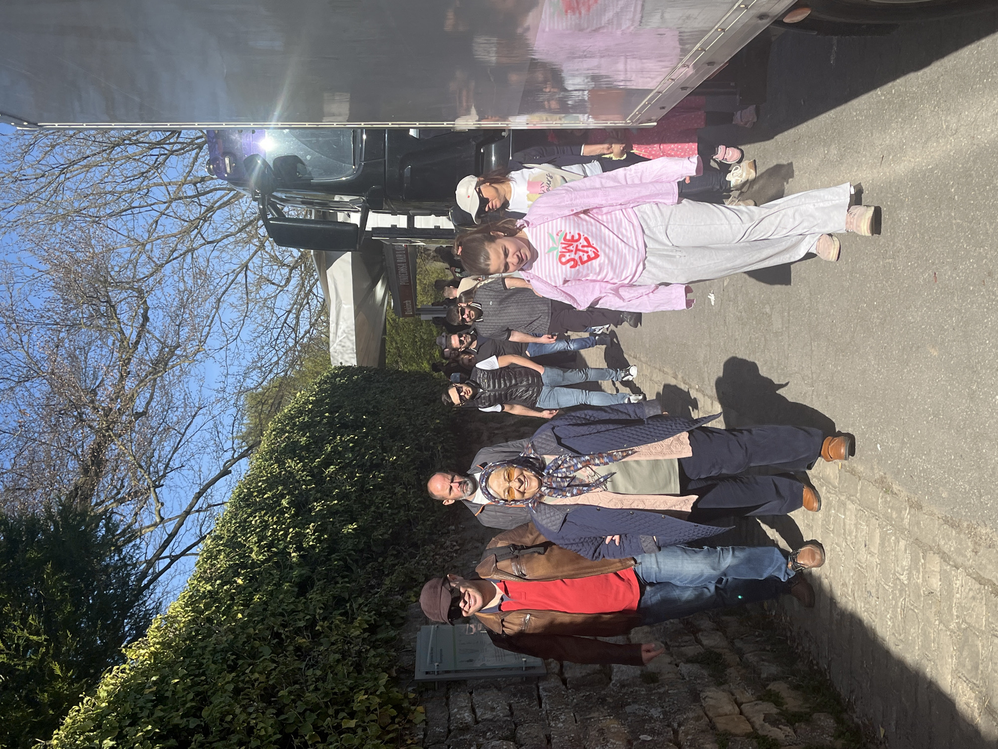 A medium shot depicts a group of eight people, possibly tourists, posing for a photograph in a public space. They are standing in a line along a cobblestone path or road, adjacent to a tall, dark green hedge on one side and a large metallic vehicle or structure on the other, possibly a truck or tour bus. The sky is visible in the upper left, indicating a clear, sunny day.
The individuals are diverse in age and attire. The person in the foreground is wearing a brown jacket, red shirt, jeans, and a cap. The next person is wearing a patterned headscarf and a blue coat or jacket. Further down the line, individuals are dressed in more casual clothing, including jeans, t-shirts, and light jackets.
The specific location in Luxembourg, Luxembourg, cannot be determined without additional context or landmarks. The presence of a tour bus or vehicle and a group of people suggests a tourist spot.