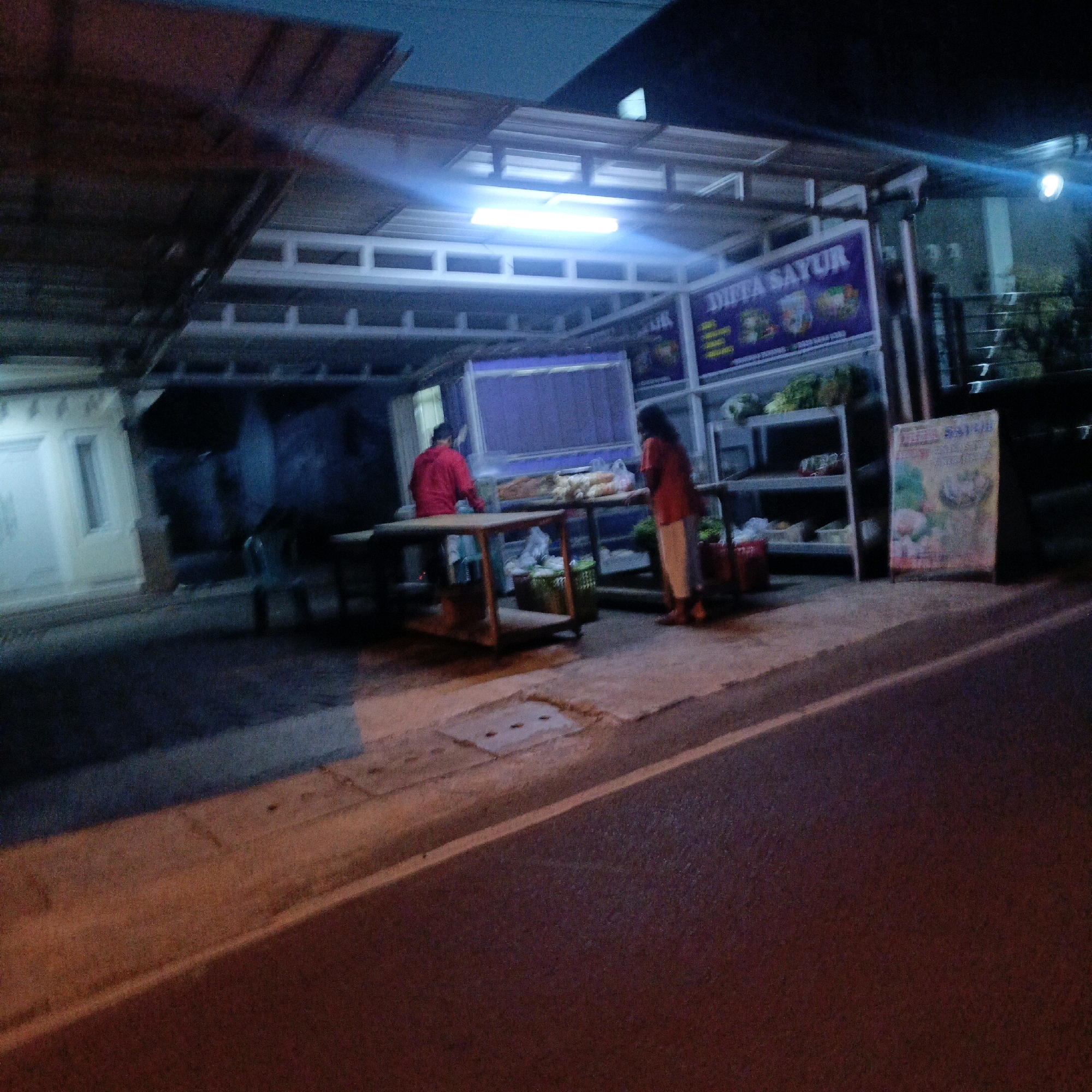 The image depicts a street-side market stall in Salatiga, Indonesia, at night. A person wearing a red jacket is standing behind a table, and another person in an orange shirt and white pants is facing away from the camera, interacting with the stall. The stall is illuminated by a bright fluorescent light overhead and has shelves filled with produce. A purple banner with text, possibly advertising "DIFA SAYUR," is visible on the stall. A sign with colorful illustrations of food is placed on the ground in front of the stall. The street in the foreground is paved and has a white line marking. The overall impression is of a humble, local business operating in a dimly lit urban environment.