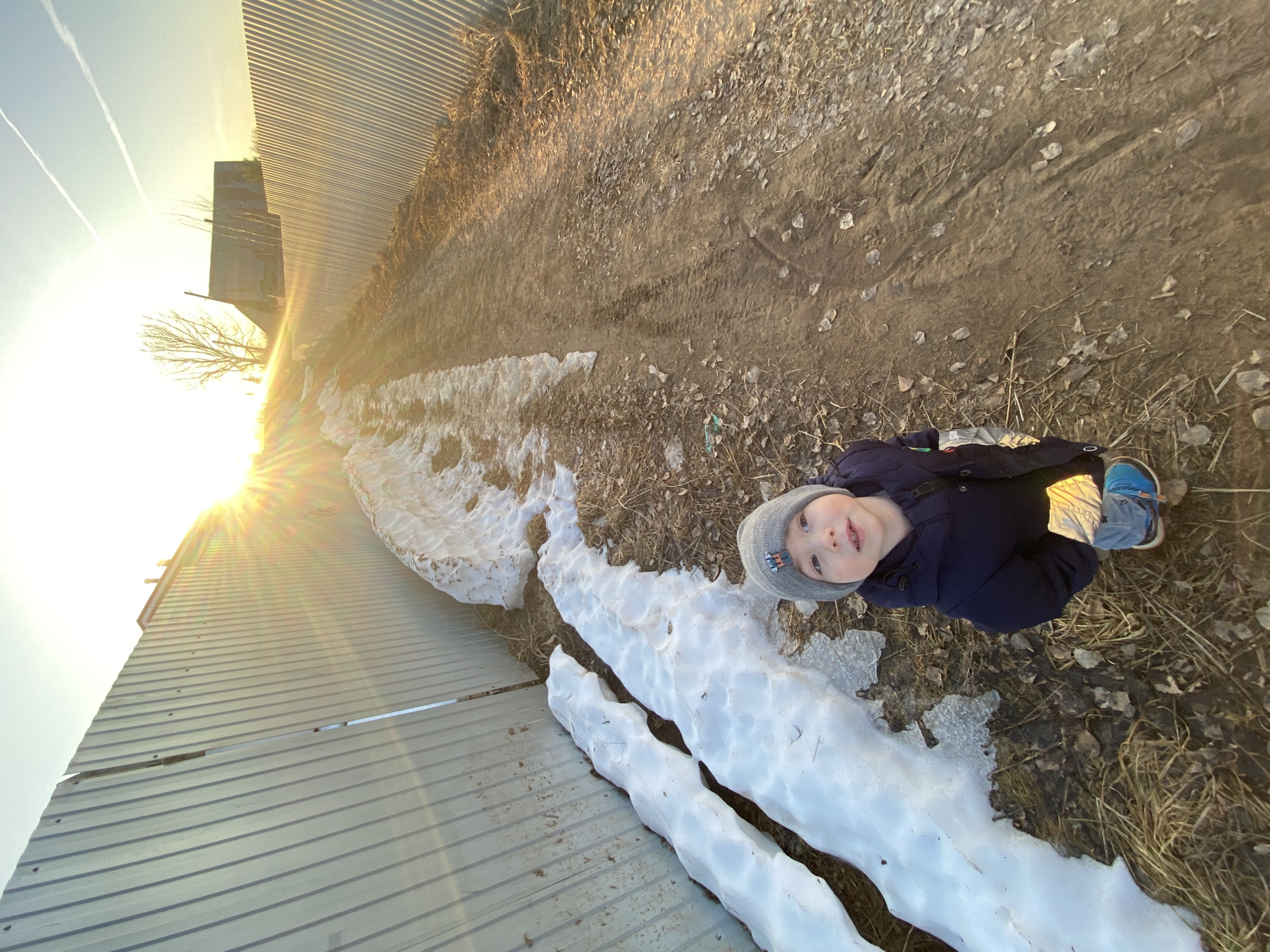 A young child stands near a patch of snow along a dirt path, adjacent to a corrugated metal fence and dried vegetation. The location is Ольховка, Russia. The sun is low in the sky, casting a strong glare.