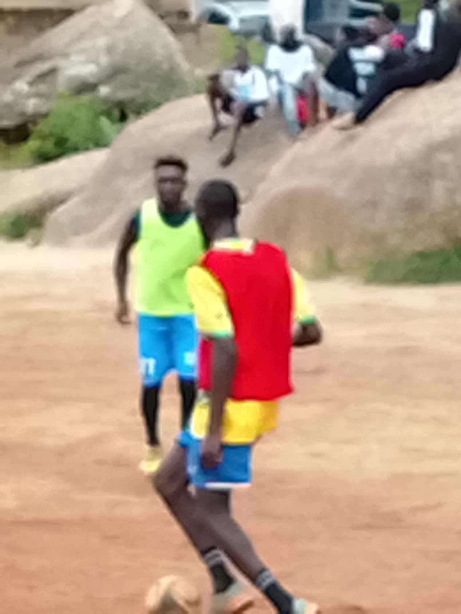 The image shows a group of people, likely young men, playing soccer on a dirt field. Two individuals are prominently featured in the foreground. One, on the left, wears a bright green vest over a dark shirt and blue shorts with white lettering. The other, on the right and facing away from the camera, wears a red vest over a yellow shirt and blue shorts. Both have dark skin and appear to be in motion, suggesting they are actively engaged in the game. A soccer ball is visible near the feet of the person on the right. In the background, other people are seated or standing on and around large, light-colored rocks. The environment appears to be a somewhat arid, outdoor setting with dry grass and sparse vegetation. The overall image quality is slightly blurred, giving it a candid feel. The specific location within "Unknown, Unknown" is not discernible from the visual information.