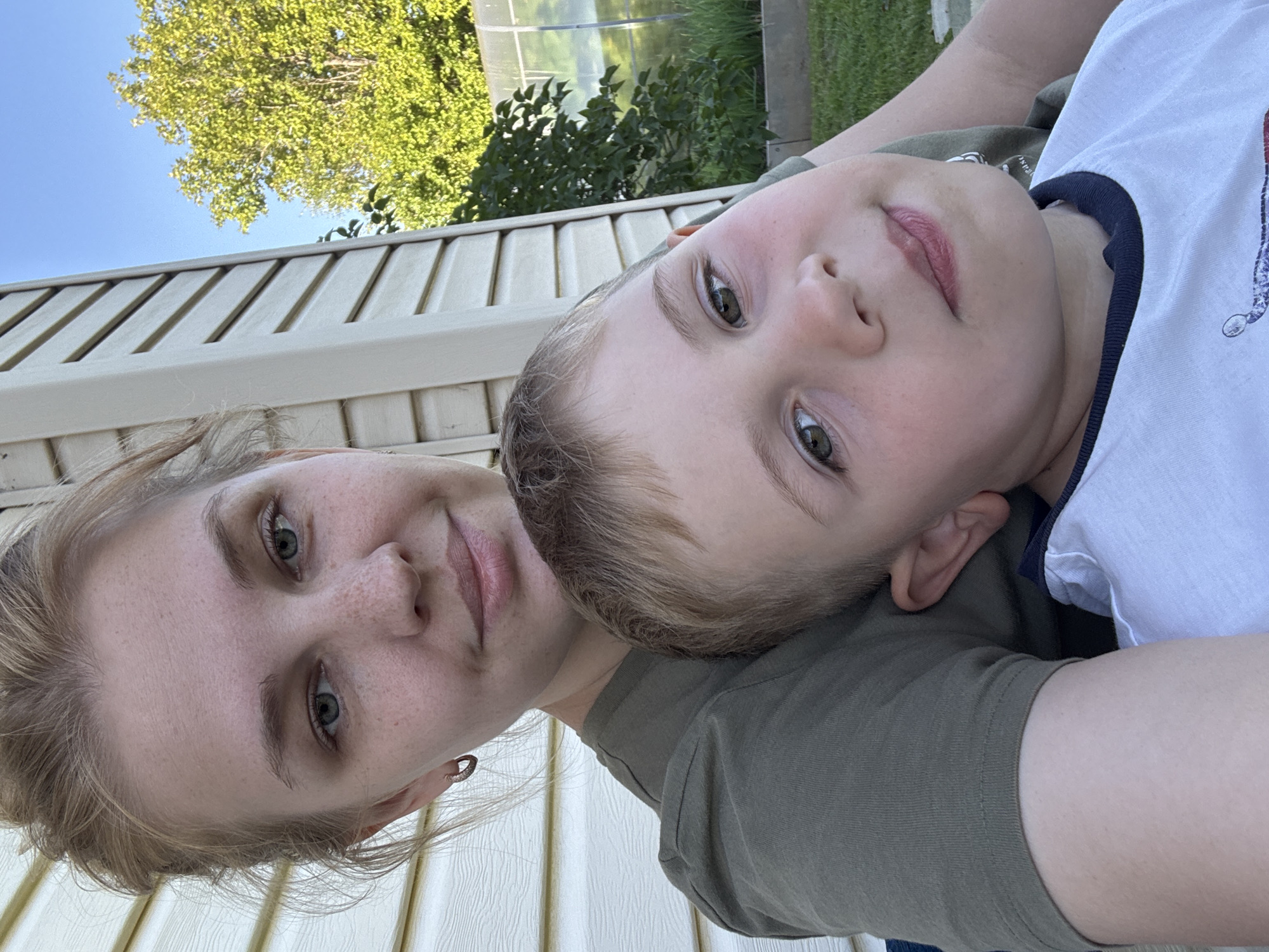 A portrait shows a young woman with blonde hair pulled back in a bun, wearing a green t-shirt and a small hoop earring. She is looking towards the camera, and her face is slightly tilted. Freckles are visible on her nose and cheeks. Beside her, a young boy with blonde hair is leaning his head on her shoulder, looking towards the camera with his eyes open. He is wearing a white t-shirt with a navy collar. They are outdoors, with a beige siding house in the background, and green trees and a greenhouse visible beyond that. The setting appears to be a residential area in the village of Dunilovo, Russia.