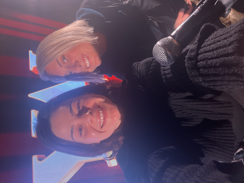 Two women smile closely at the camera. One has shoulder-length blonde hair and wears a dark jacket. The other, positioned lower, has brown hair and wears a black sweater. A microphone is visible, held near the black sweater. A large, illuminated sign with red, orange, and blue colors is behind them, with an "A" prominently visible. The lighting is bright, highlighting their faces and teeth.