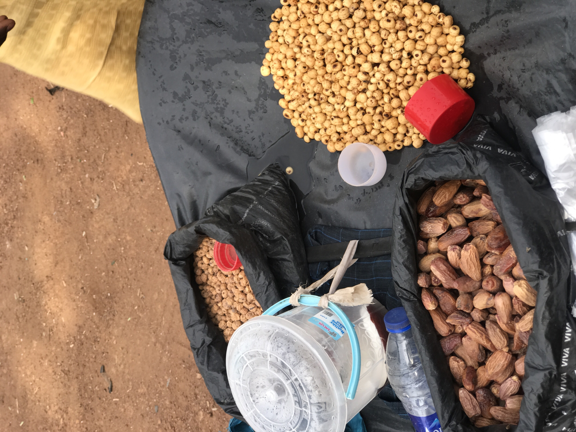 This overhead shot depicts a market stall or roadside vendor's display in Galadimawa Village, Nigeria, under natural daylight. The setting features reddish-brown sandy ground, with a portion of a yellow fabric or mat visible on the left. A dark gray or black tarp, which appears damp, serves as the primary display surface.
The main items for sale include a substantial pile of light brown, small, rounded-oval nuts, likely tiger nuts or chufa, spread across the upper right section of the tarp. Nearby, an overturned red plastic cup and a small, clear plastic measuring cup are present.
To the right, a black plastic bag is filled with numerous dried dates. The bag has "VIVA VIVA" printed repeatedly on its visible surface. Another black plastic bag, also containing light brown nuts similar to the main pile, is positioned in the lower-left.
Additional objects contributing to the scene include a clear plastic bucket with a light blue handle, appearing to hold liquid, and bearing a label that partially shows "BALWYN". Fabric strips are tied to the bucket's handle. A plastic water bottle with a blue cap, with the brand name "AQUA" visible, rests beside the bag of dates. A stack of white plastic bags is also seen, presumably for packaging purchases.
No individuals are clearly visible in the image. The arrangement of goods suggests a common method of displaying produce for sale in a local market environment.