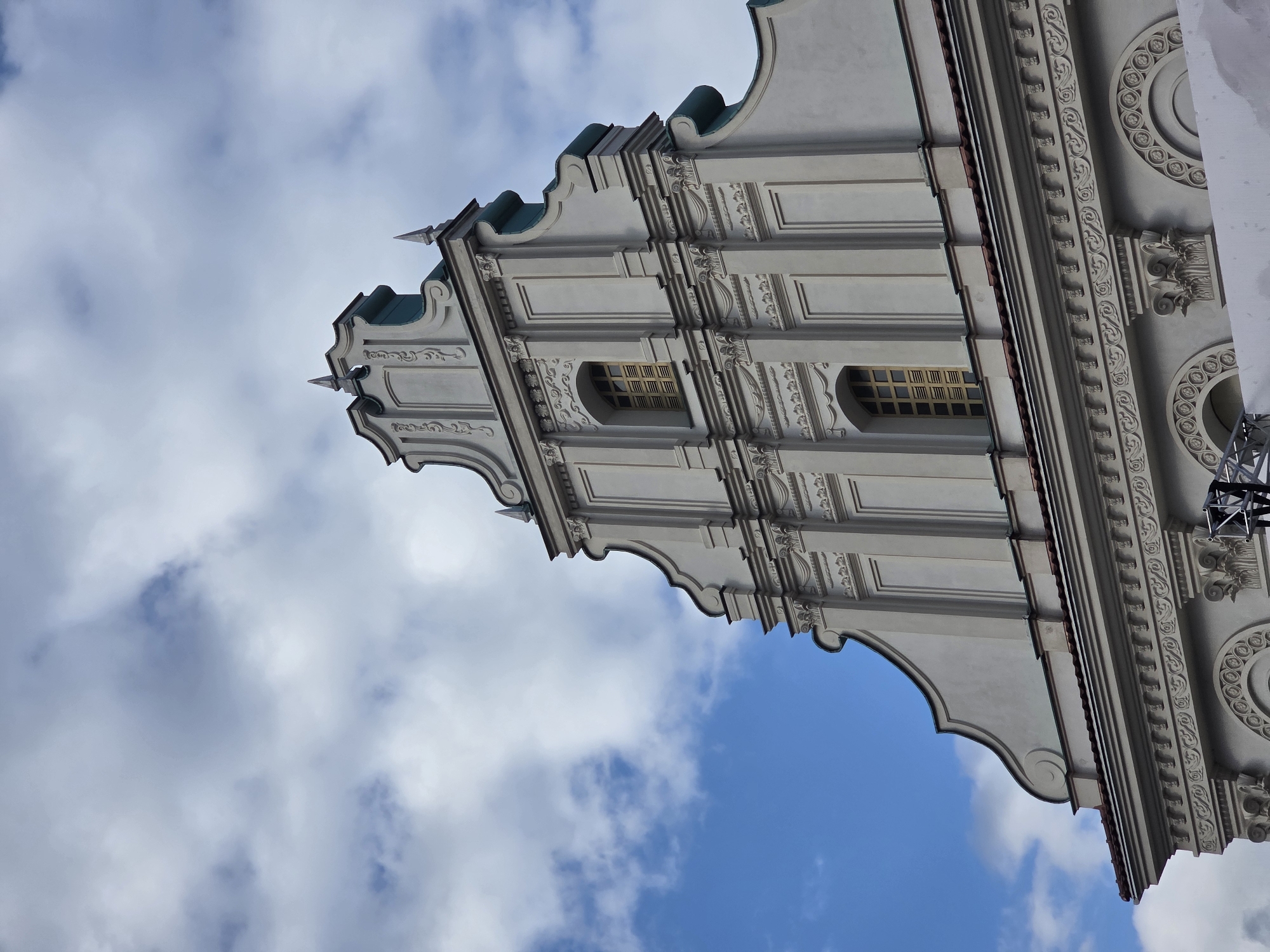 The image shows the upper facade of a classical building against a sky with scattered clouds. The building features a stepped gable with decorative molding, pilasters, and two rectangular windows with wooden shutters. Below the gable, a cornice runs horizontally, adorned with intricate relief work. Further down, another architectural detail includes rounded niches and column capitals. The color palette is dominated by white and grey for the building, with hints of teal on the roofline and a reddish-brown for the roof tiles. The sky is a mix of blue and white clouds, suggesting it is daytime. No people or specific activities are visible.