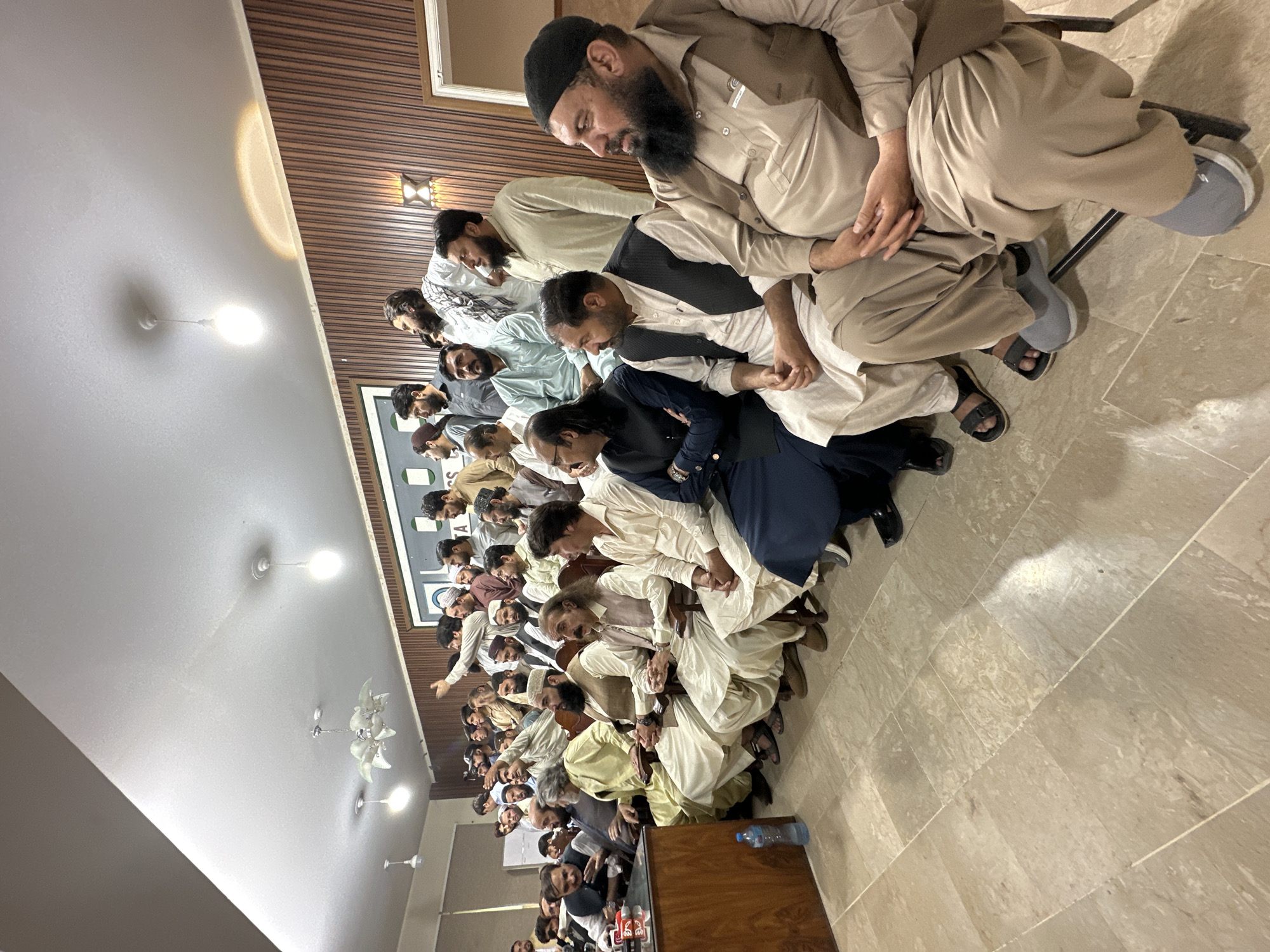 A wide-angle, slightly rotated indoor shot shows a large group of men sitting on chairs in rows. The men are dressed in traditional Pakistani clothing, including shalwar kameez and waistcoats. Many men wear prayer caps or turbans. They are seated facing a wooden lectern. A few people are standing in the foreground by the lectern. The room has a tiled floor, a white ceiling with lights and ceiling fans, and wood-paneled walls. The location is Quetta City Tehsil, Pakistan.