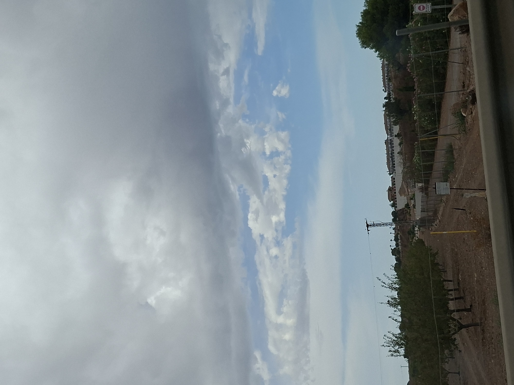 The image captures a cloudy sky over a landscape in Murcia, Spain. In the foreground, a dirt road runs alongside a row of trees, suggesting an orchard or park. A fence encloses the area, and a yellow pole stands by the roadside. In the background, buildings are visible on a hill, and a power pole stands on the left. The overall scene depicts a rural or semi-urban environment under overcast skies.