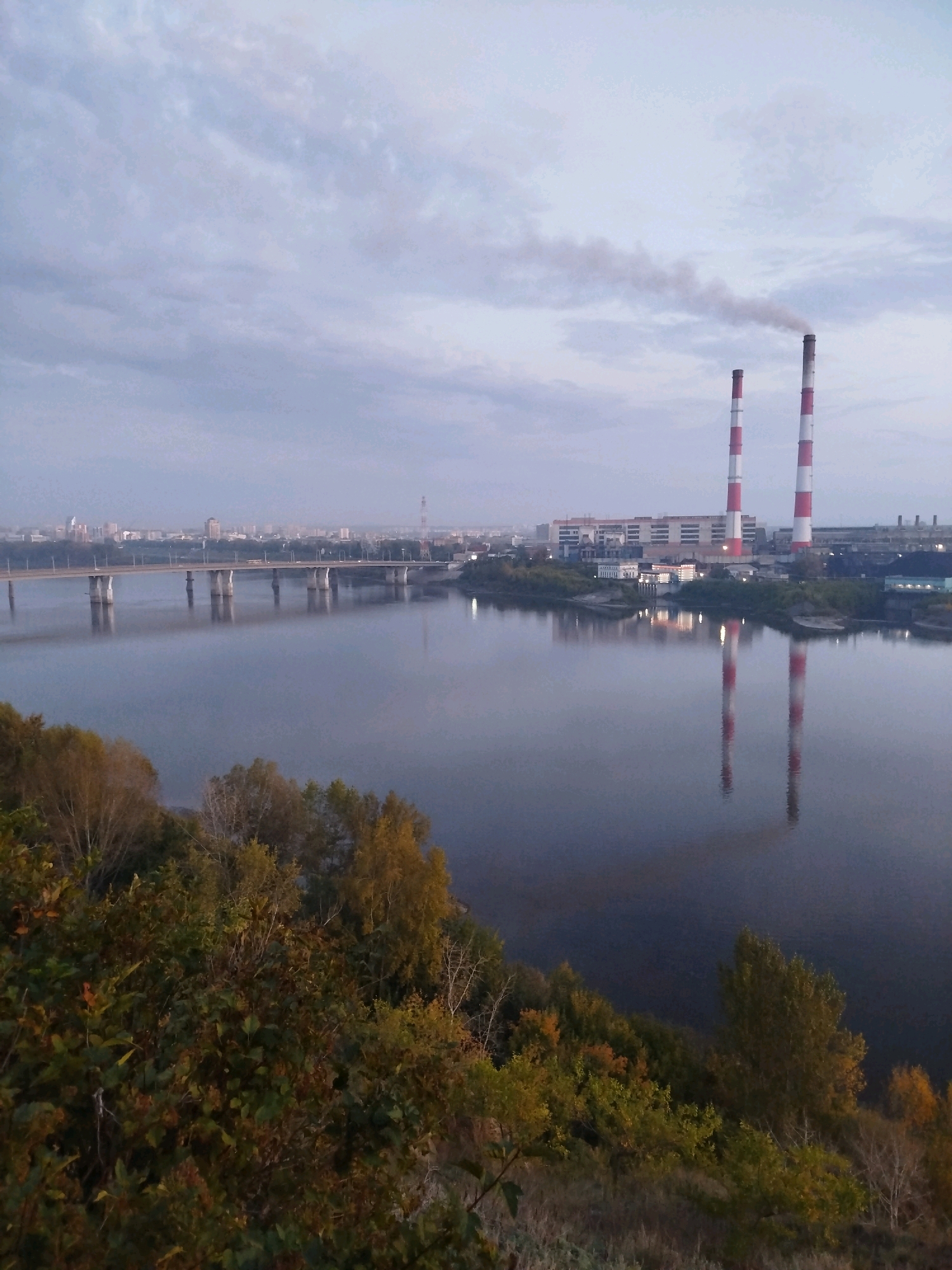 A wide shot captures an industrial scene at dusk in Kemerovo, Russia, dominated by a large river reflecting the sky and the environment. On the far bank, two prominent red and white striped smokestacks rise, emitting a plume of grey smoke into the overcast sky. Adjacent to the smokestacks is a long, low industrial building complex. In the midground, a wide concrete bridge with multiple arches spans the river, with a line of streetlights visible along its length. Beyond the bridge, the distant city skyline is faintly visible, characterized by low-rise buildings and a solitary tall tower. The foreground is a steep embankment covered with dense, autumnal foliage in shades of green, yellow, and brown, suggesting a transition into cooler weather. The water's surface is calm, creating clear reflections of the bridge and the industrial structures. The overall mood is somber and industrial, typical of a city with significant manufacturing presence.