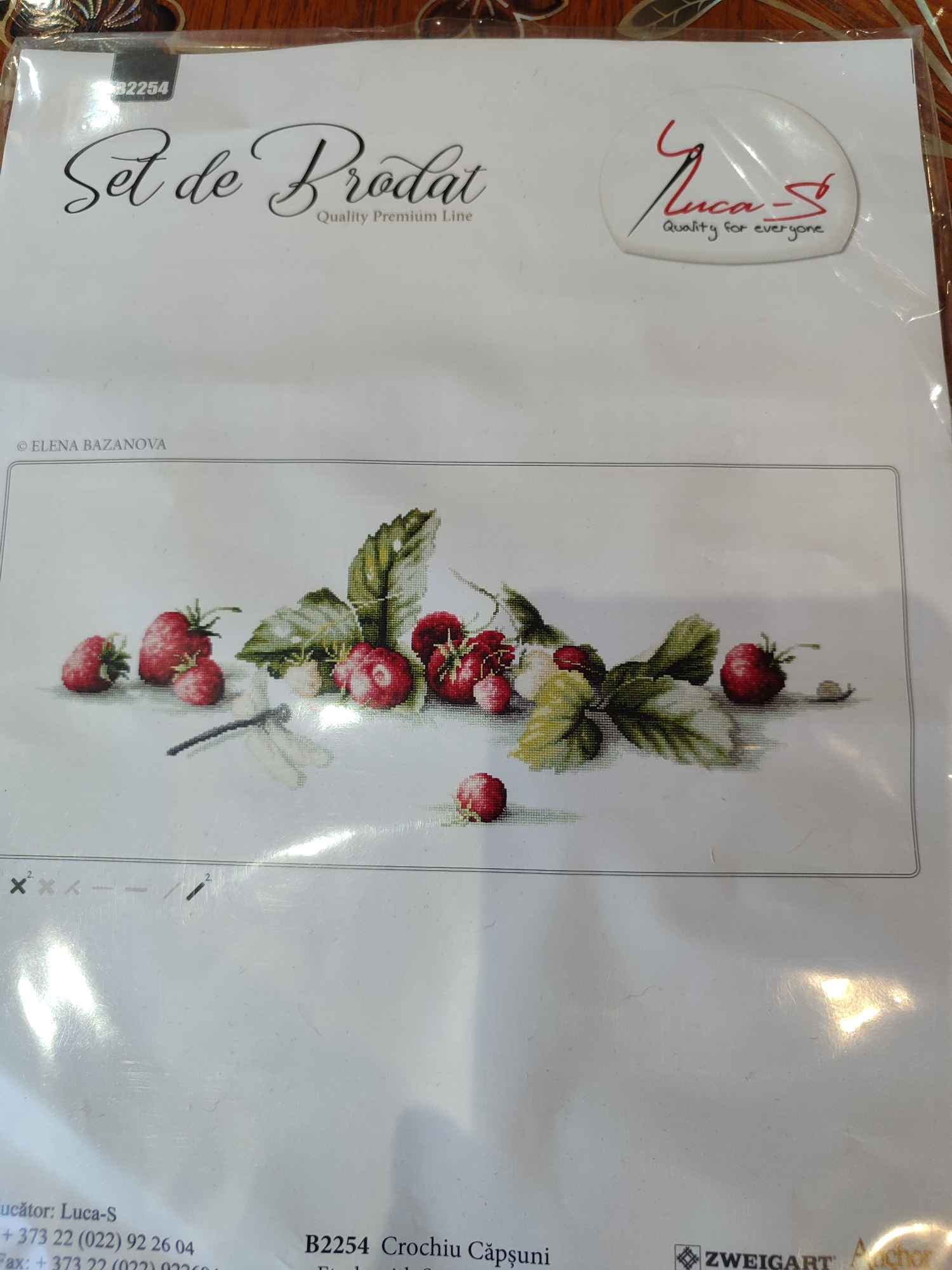 The image shows the packaging of a cross-stitch kit. The packaging features a printed image of strawberries and a dragonfly. Text on the packaging includes "Set de Prodat", "Quality Premium Line", "B2254 Crochiu Căpșuni", and a logo that says "Luca-S Quality for everyone". There is also a copyright notice "© ELENA BAZANOVA". The bottom of the packaging includes contact information for "Luca-S" and a logo for "ZWEIGART Anchor". The image is taken from a top-down perspective, with the packaging lying flat on a wooden surface. There are no people or any indication of the city of Tuapse, Russia.