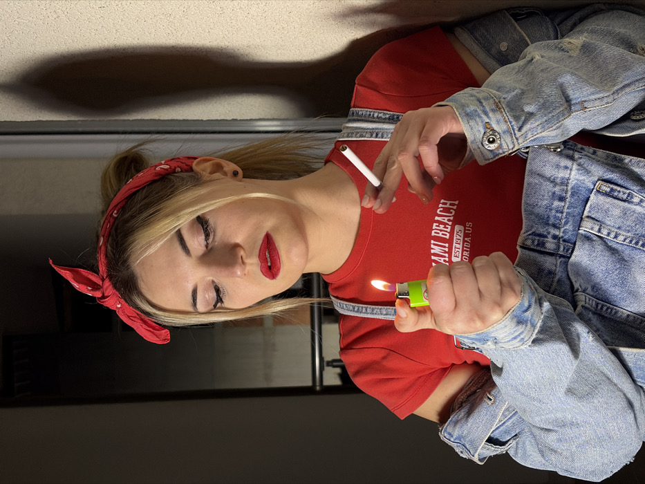 A young adult female is depicted indoors in Budva, Montenegro, engaged in the act of lighting a cigarette. She has blonde hair pulled back, adorned with a red bandana patterned with white elements. Her facial features include dark eyeliner and prominent red lipstick. She wears a red t-shirt with "MIAMI BEACH EST. 1975 FLORIDA.US" printed in white, blue denim overalls, and a light blue denim jacket draped over her shoulders. In her left hand, she holds an activated green lighter, producing a visible flame. Her right hand holds a white, unlit cigarette positioned near the flame. Her gaze is directed downwards, focused on the lighter and cigarette. The background on the left is dark, suggesting an interior space or doorway, while the right side features a light-colored, textured wall with a distinct, elongated shadow cast upon it.