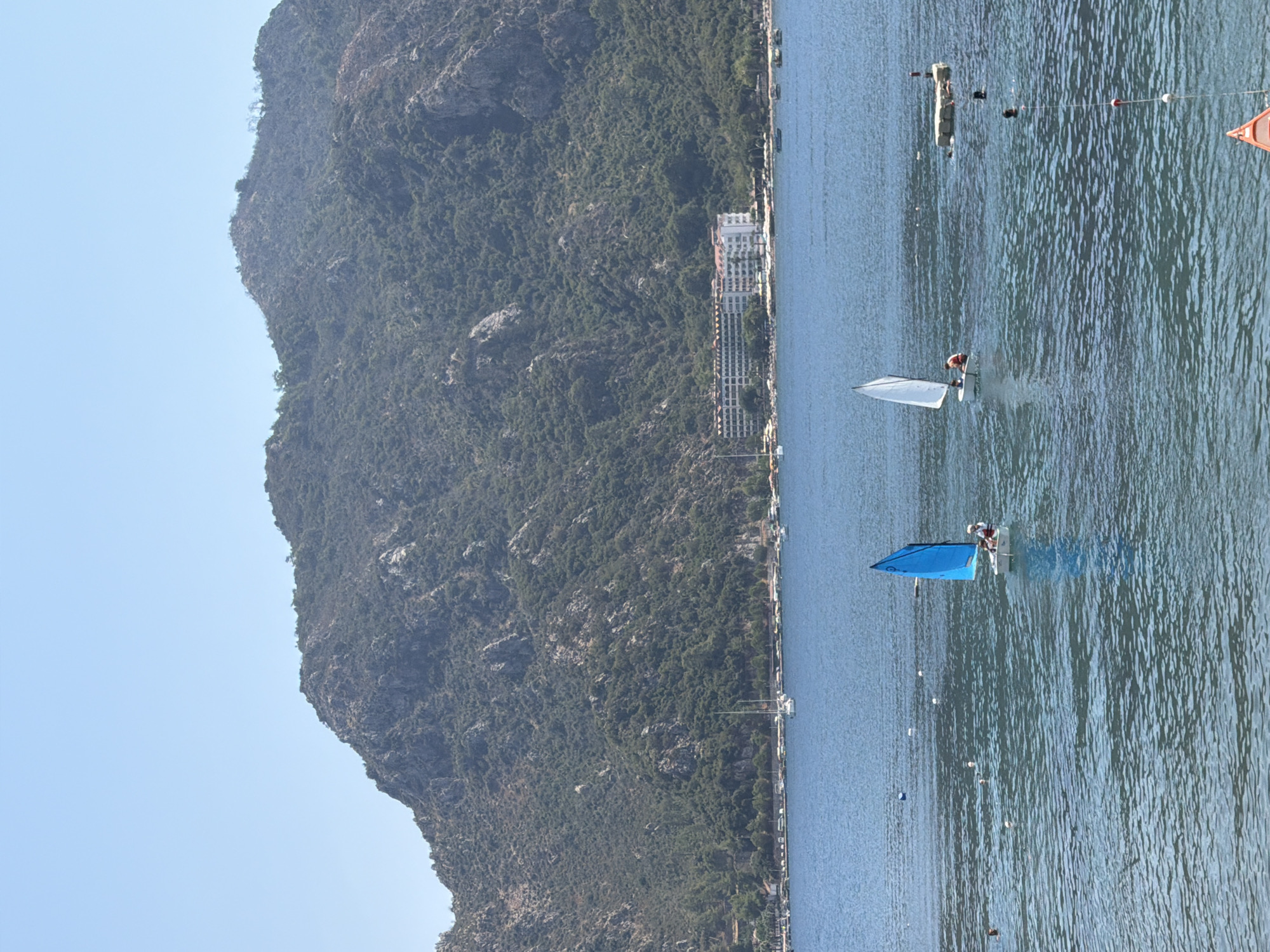 The image depicts a scenic view of Marmaris, Turkey, on a clear, sunny day. In the foreground, the calm, blue waters of the Aegean Sea are visible, with two small sailboats actively sailing. The sailboat on the left has a bright blue sail, and the one on the right has a white sail. Both sailboats appear to have at least one person aboard, engaged in sailing. Several other people are swimming or enjoying the water in various parts of the sea.
In the mid-ground, the coastline of Marmaris is lined with a strip of hotels and buildings, with lush green trees and vegetation dominating the landscape. The buildings appear modern, with several stories and balconies. Further in the background, a large, imposing mountain covered in dense green foliage rises majestically. The mountain's rocky terrain is visible in places, adding to the dramatic backdrop. The sky above is a clear, unblemished blue, indicating a warm and pleasant day. A few other boats are anchored or sailing further out in the bay, and a small floating platform with a red marker is also present. The overall impression is a tranquil and inviting coastal resort scene.