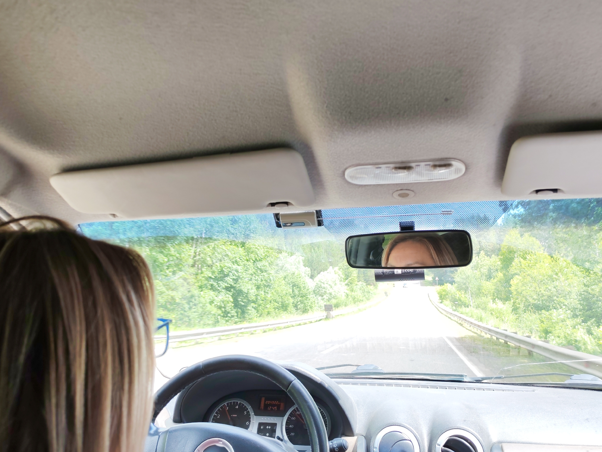 The image shows the interior of a car, looking forward through the windshield. The driver's head and shoulders are visible in the foreground on the left, with blonde hair. The dashboard is in the lower part of the frame, including a steering wheel and instrument cluster displaying "204922 km" and "12:45". A rearview mirror is mounted on the windshield, reflecting the driver's face, specifically her eyes and forehead. The road ahead is visible, curving to the right and lined with trees and vegetation. The sky is bright, suggesting daytime. This scene is not specifically identifiable as being in Buranovo, Russia; it depicts a common car interior and a road in a natural setting.