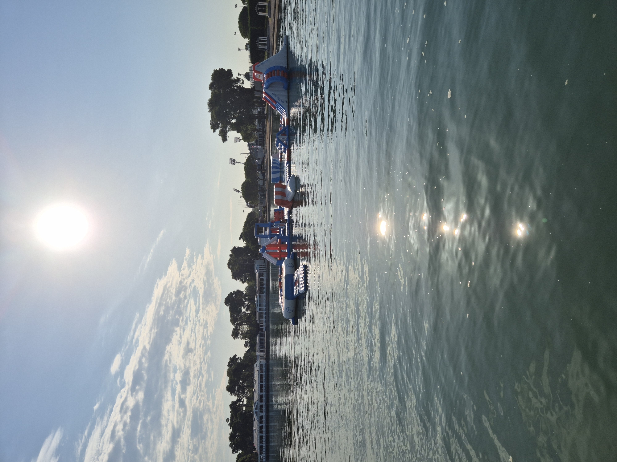 This is a landscape photograph taken in Umag, Croatia, showing an inflatable water park set up on a body of water. The sun is high in the sky, casting a bright glare on the water's surface and creating reflections. In the background, there is a shoreline with trees, buildings, and what appear to be sports stadium lights. The water park consists of various inflatable structures, including slides and climbing elements, in blue, white, and orange. The water is calm with gentle ripples, and some small white objects, possibly foam or debris, are floating on the surface. The scene suggests a recreational area, likely during daylight hours.