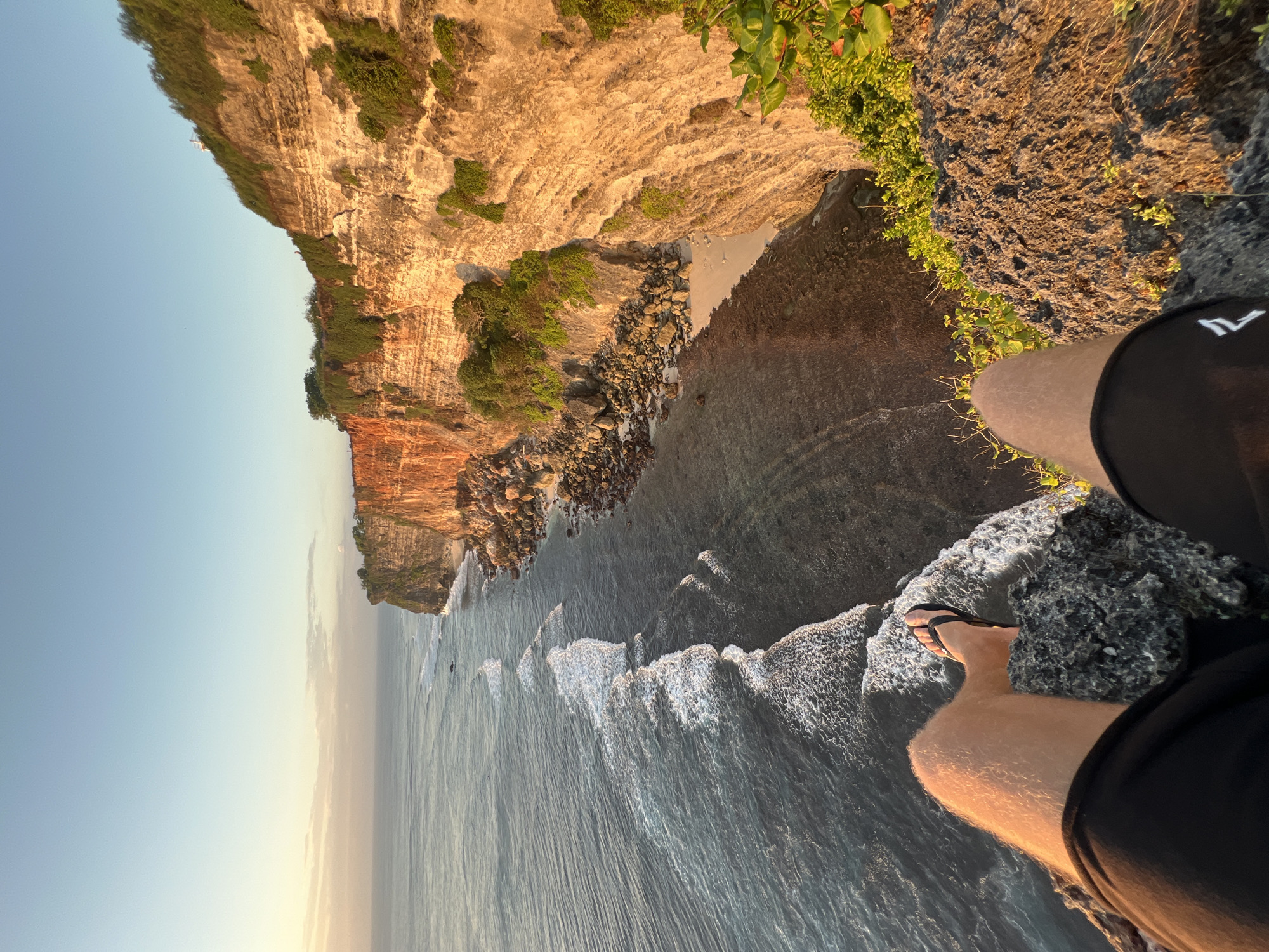 The image depicts a person's legs and feet, wearing black shorts and black flip-flops, sitting on the edge of a rocky cliff overlooking the ocean. The sun is low in the sky, casting a warm golden light on the cliff face and the water. The ocean below is calm with gentle waves lapping at the shore. The cliff is covered in lush green vegetation. In the distance, the coastline stretches out, with more cliffs and vegetation visible. The scene appears to be a scenic viewpoint in a coastal area. Based on the context of Kuta Selatan, Indonesia, this is likely a view of Uluwatu cliff.