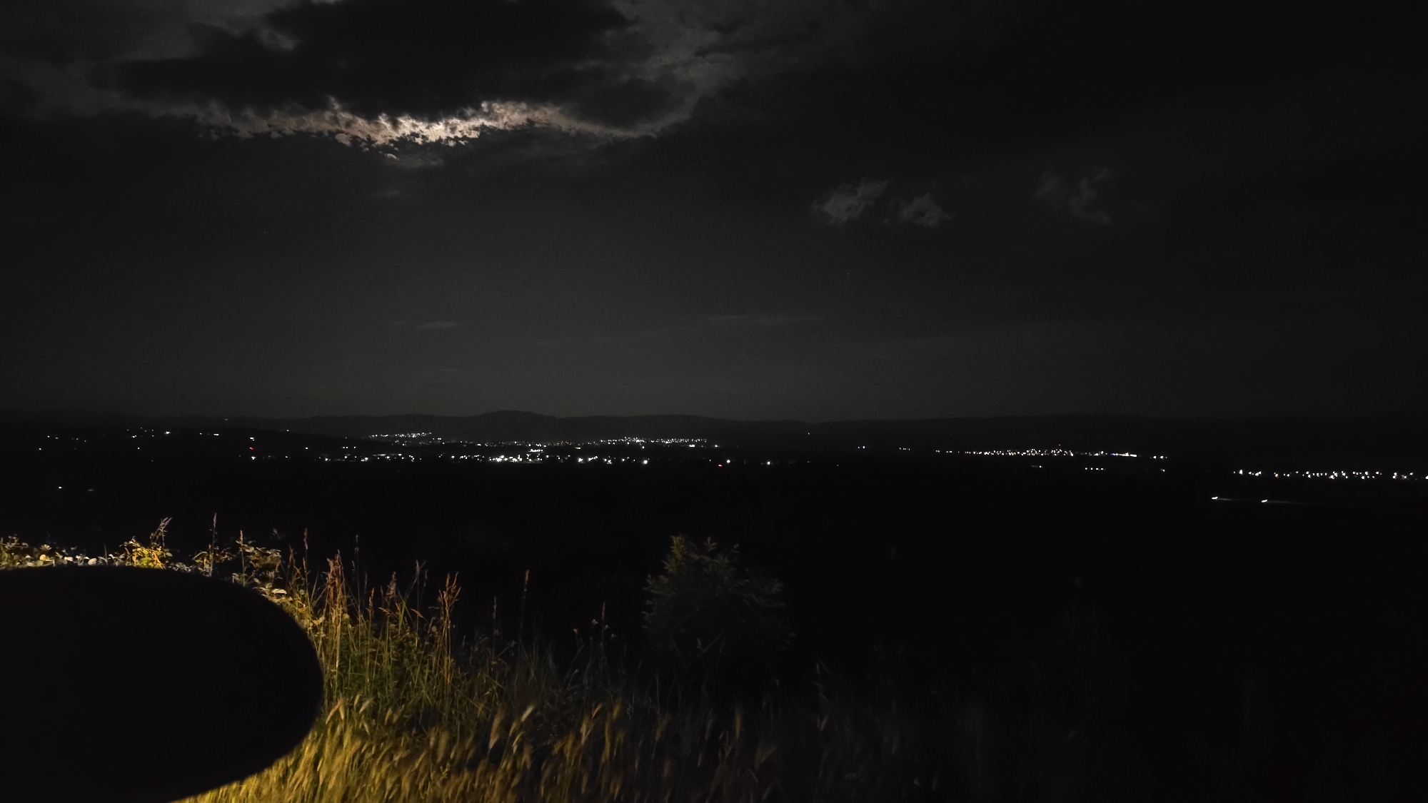 The image shows a night scene, likely a landscape view from a high vantage point in Kran, Bulgaria.
**Environment:** The scene is dark, with a night sky containing clouds. The moon is partially visible through a break in the clouds, casting a dim light. In the distance, a city or town is visible, indicated by clusters of lights. The foreground features vegetation, likely tall grasses or reeds.
**Objects:** Distant city lights, moon obscured by clouds, tall grasses. A dark, circular object is in the lower left corner of the image.
**Actions:** Moonlight filtering through clouds.
**Interactions:** Light illuminating the distant city.