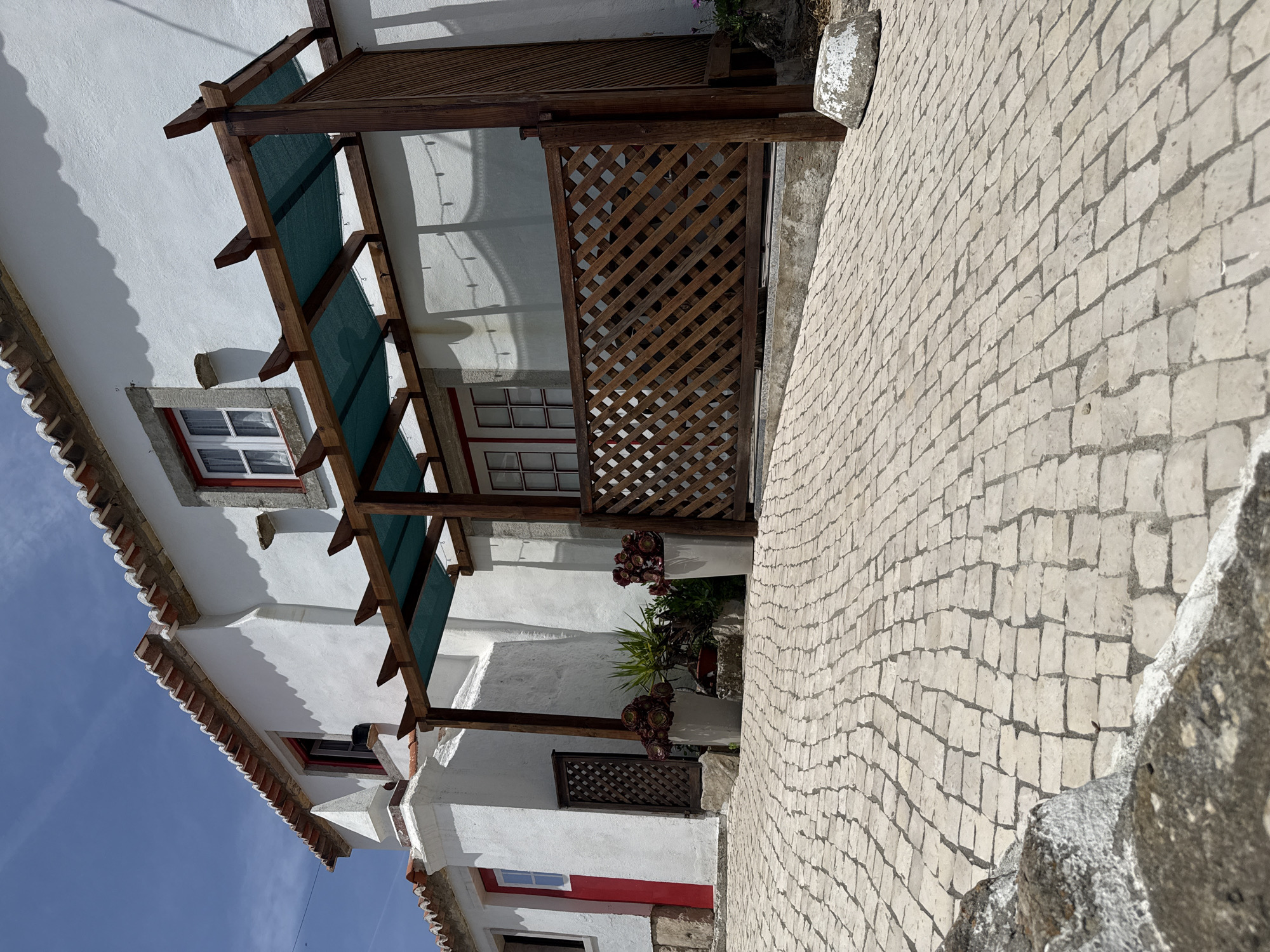 The image shows a white building with a red-tiled roof and red-trimmed windows, likely a house or a small establishment, situated on a cobbled street in the town of Colares, Portugal. A wooden pergola with a green mesh cover extends from the building, providing shade over a patio area. The patio is enclosed by a wooden lattice fence. Two large white planters with succulent plants are placed near the entrance. The street is paved with light-colored cobblestones that slope upwards. The sky is clear blue with some wispy clouds. The scene is bathed in bright sunlight, creating strong shadows. There are no people visible in the image.